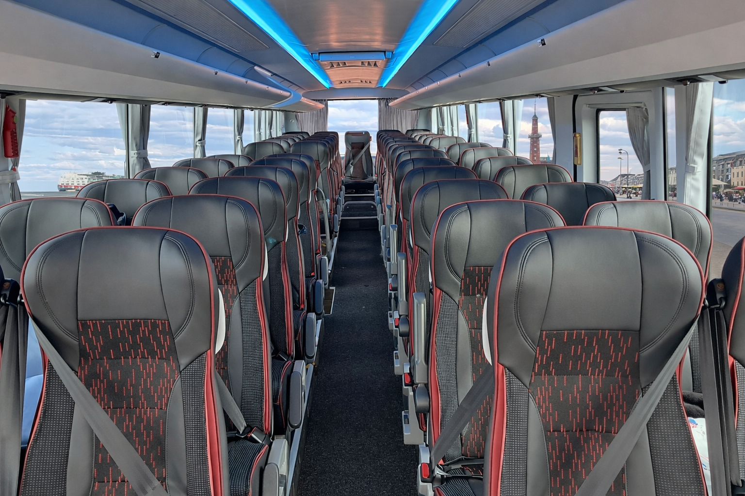 Interior of a modern bus with rows of black and red cushioned seats, large windows, and a view of waterfront and cityscape outside.