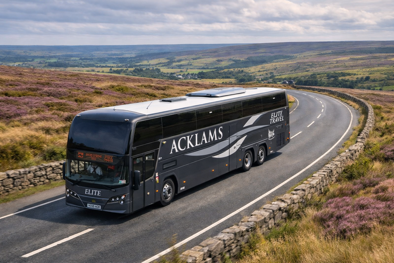 A black coach bus with 'ACKLAMS' and 'ELITE TRAVEL' written on it driving along a winding road through a rural landscape with stone walls and purple heather