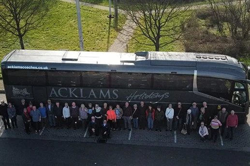 A bus with people standing in a line outside, parked next to a sidewalk and grassy area with leafless trees.