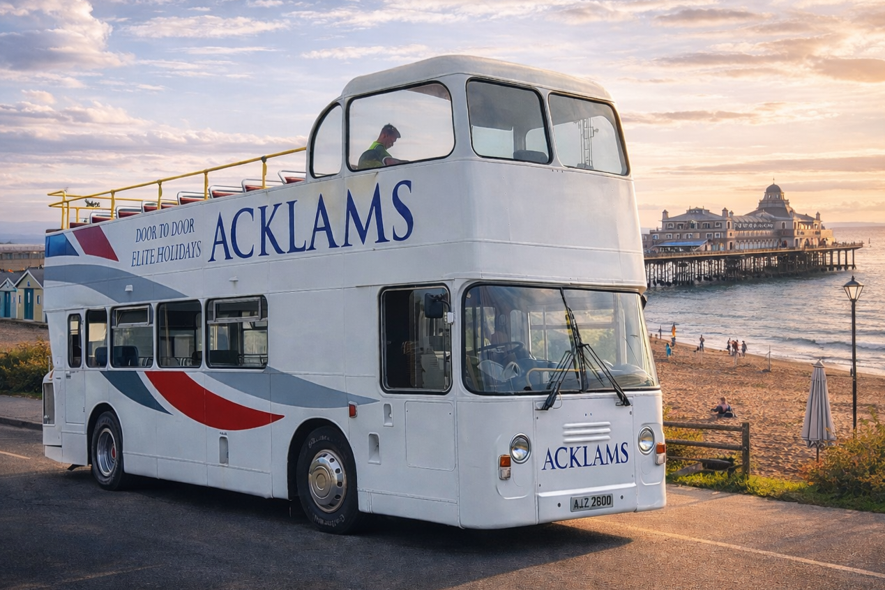Double-decker sightseeing bus with 'Acklams' branding parked near a beach at sunset, with a pier and historic building in the background.