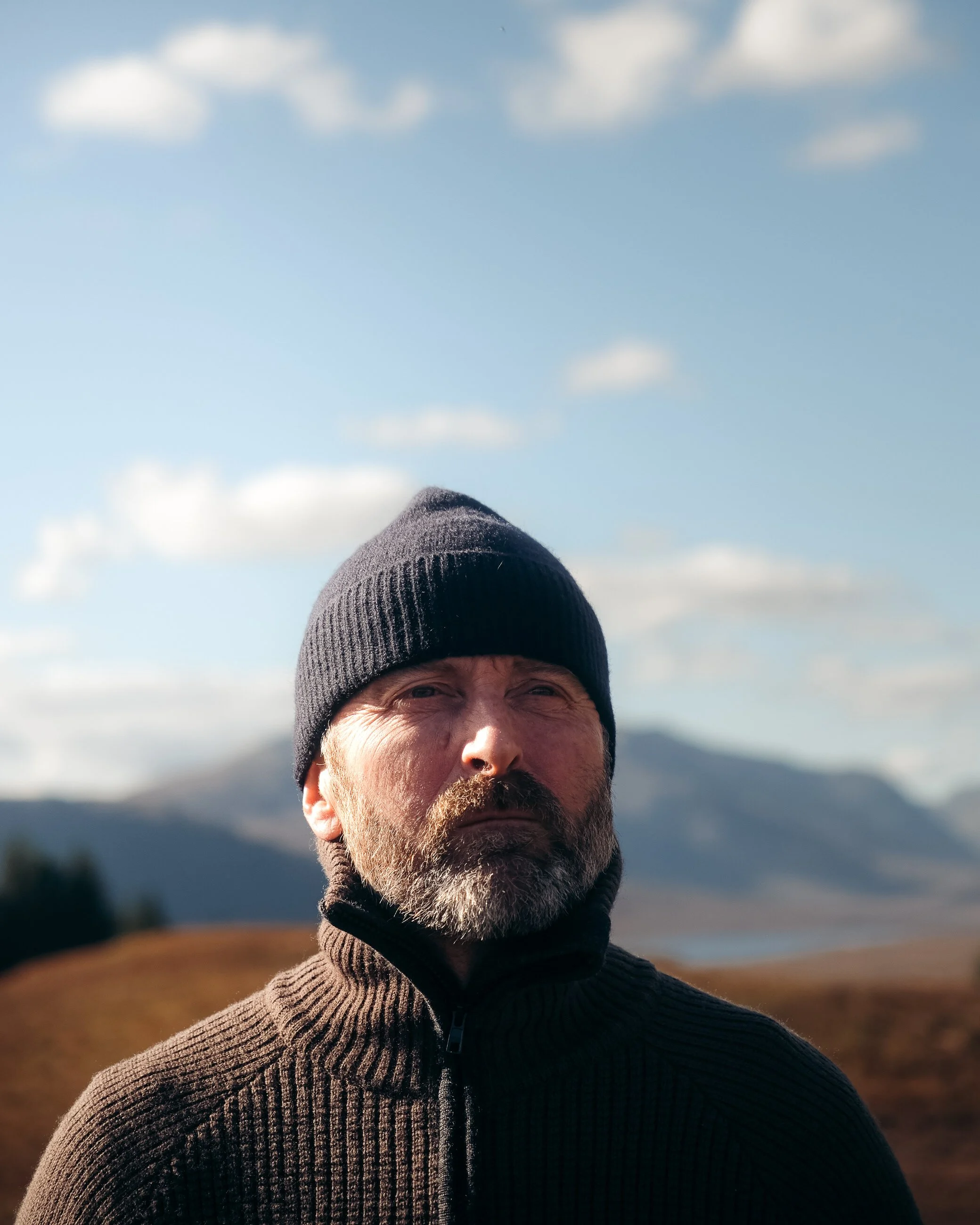 A man wearing a black beanie and black zip-up jacket standing outdoors with mountains and a partly cloudy sky in the background.