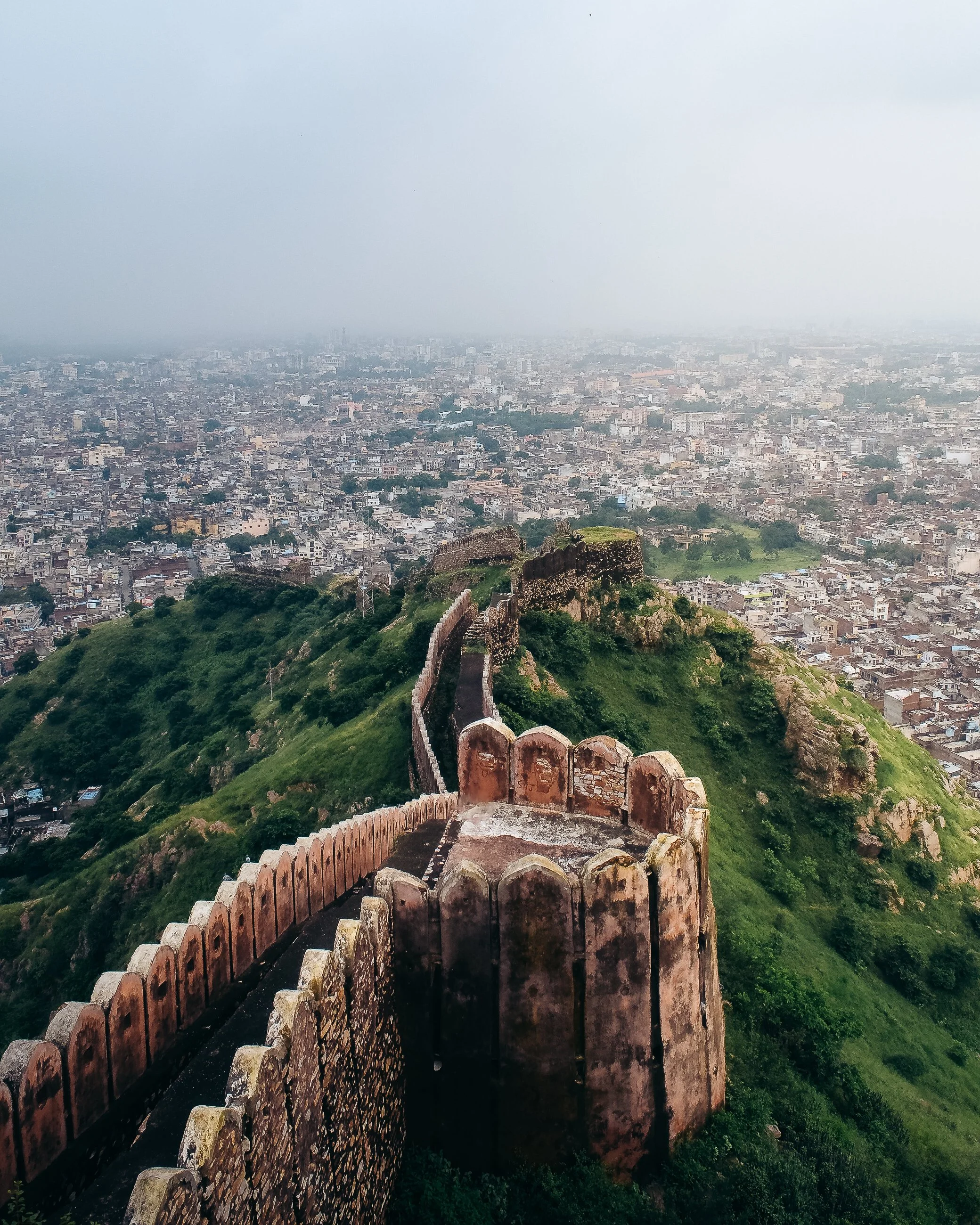 View of a historic fort or wall on a green hilltop overlooking a densely populated city with many buildings and streets, under a cloudy sky.