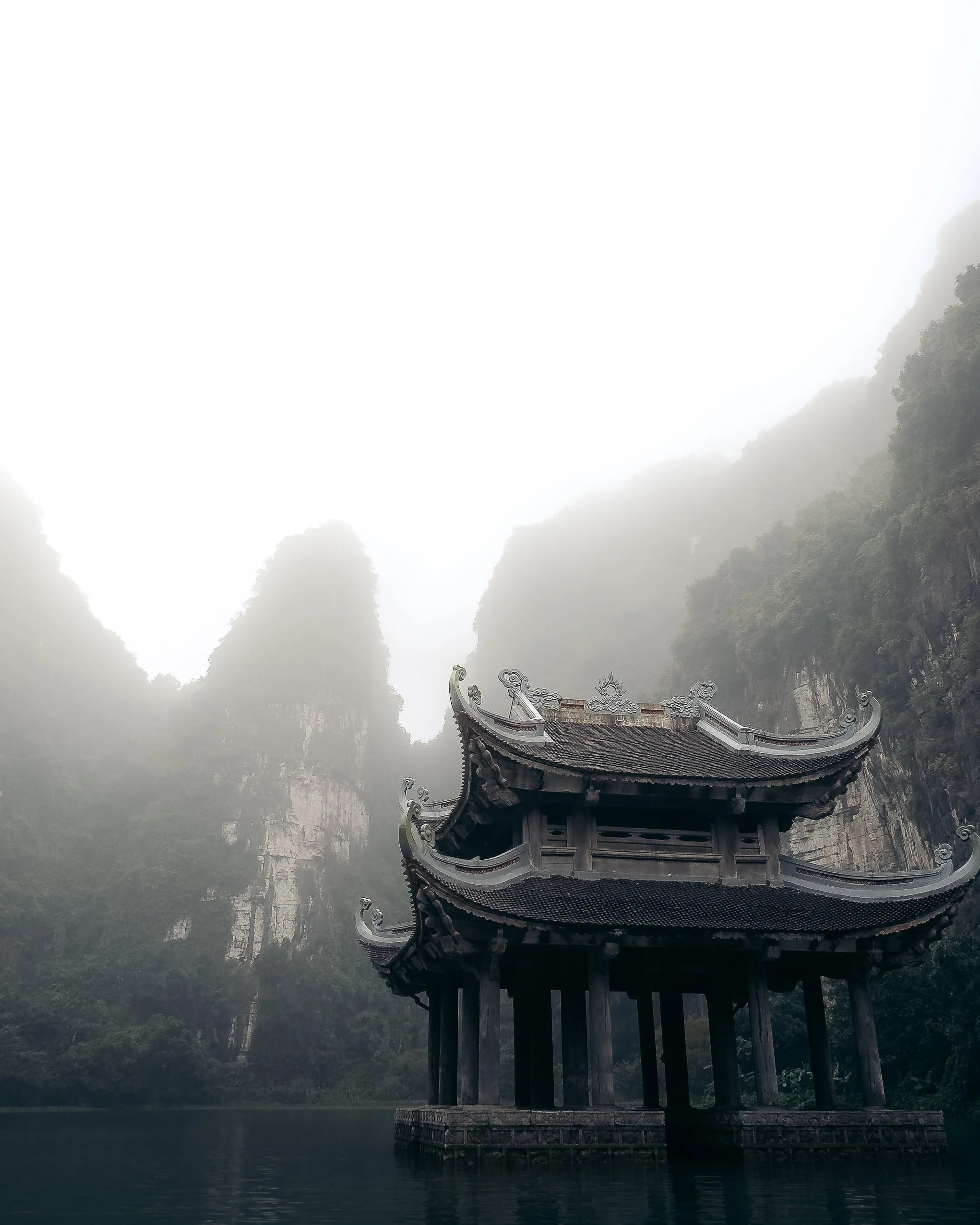 Traditional Asian-style pavilion with curved roofs, situated on water with misty mountains in the background.