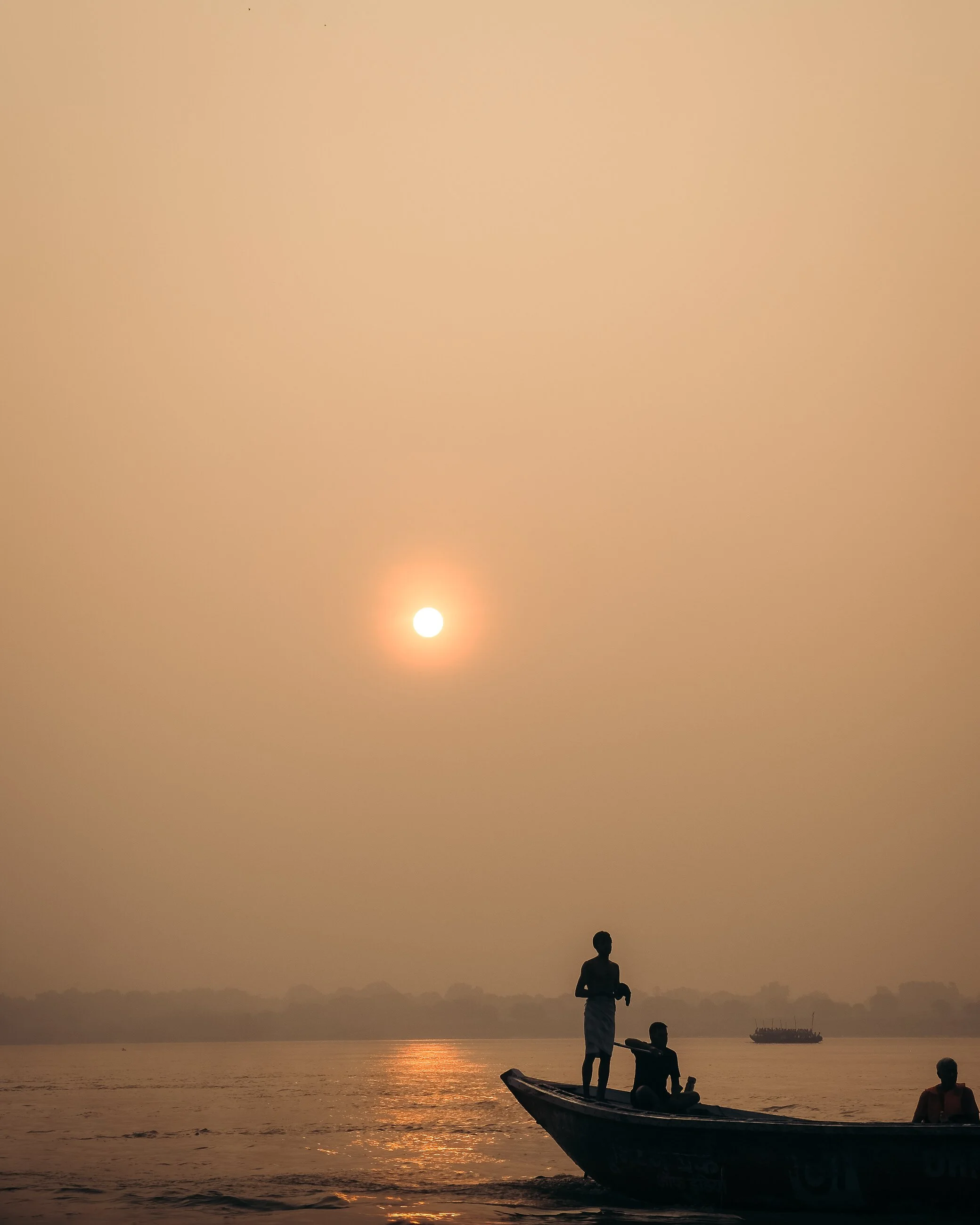 Silhouettes of people on a boat on a river during sunset or sunrise with a hazy sky and a distant boat on the water.