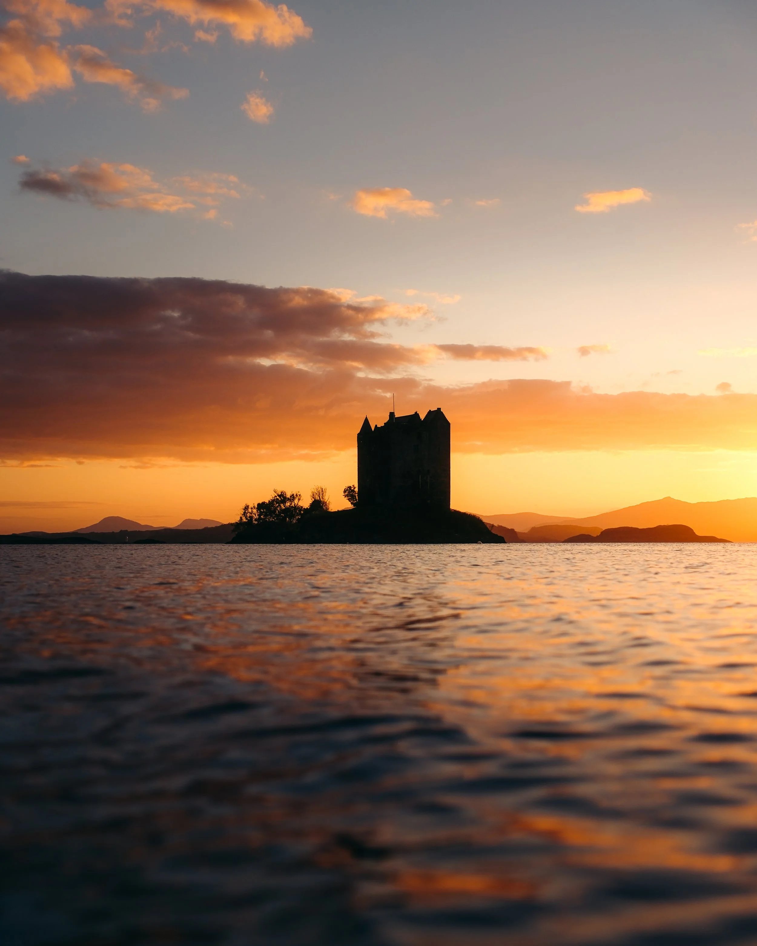 Silhouette of a castle on a small island at sunset with orange and blue sky and water in the foreground.