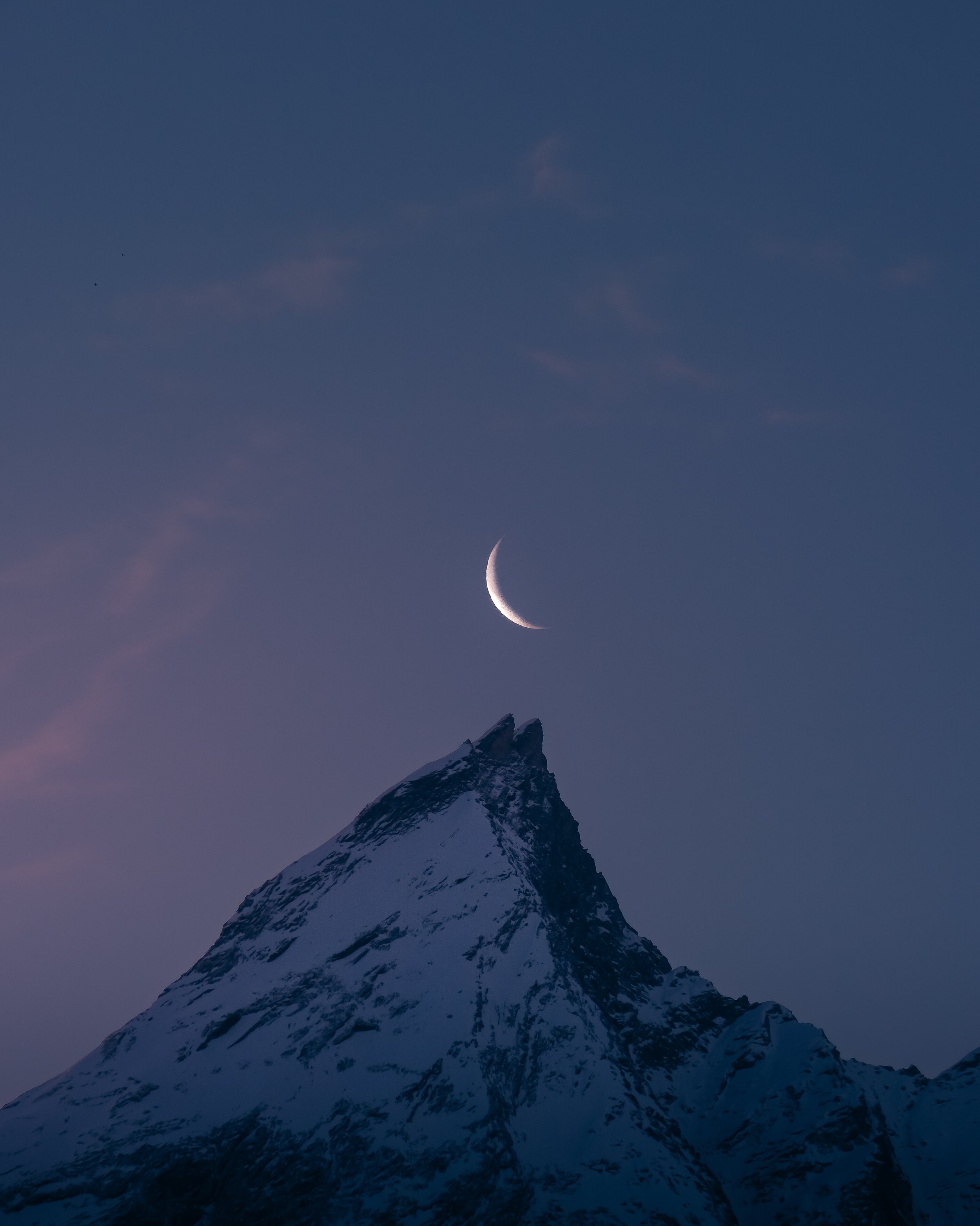 Snow-covered mountain peak with a crescent moon in the evening sky.