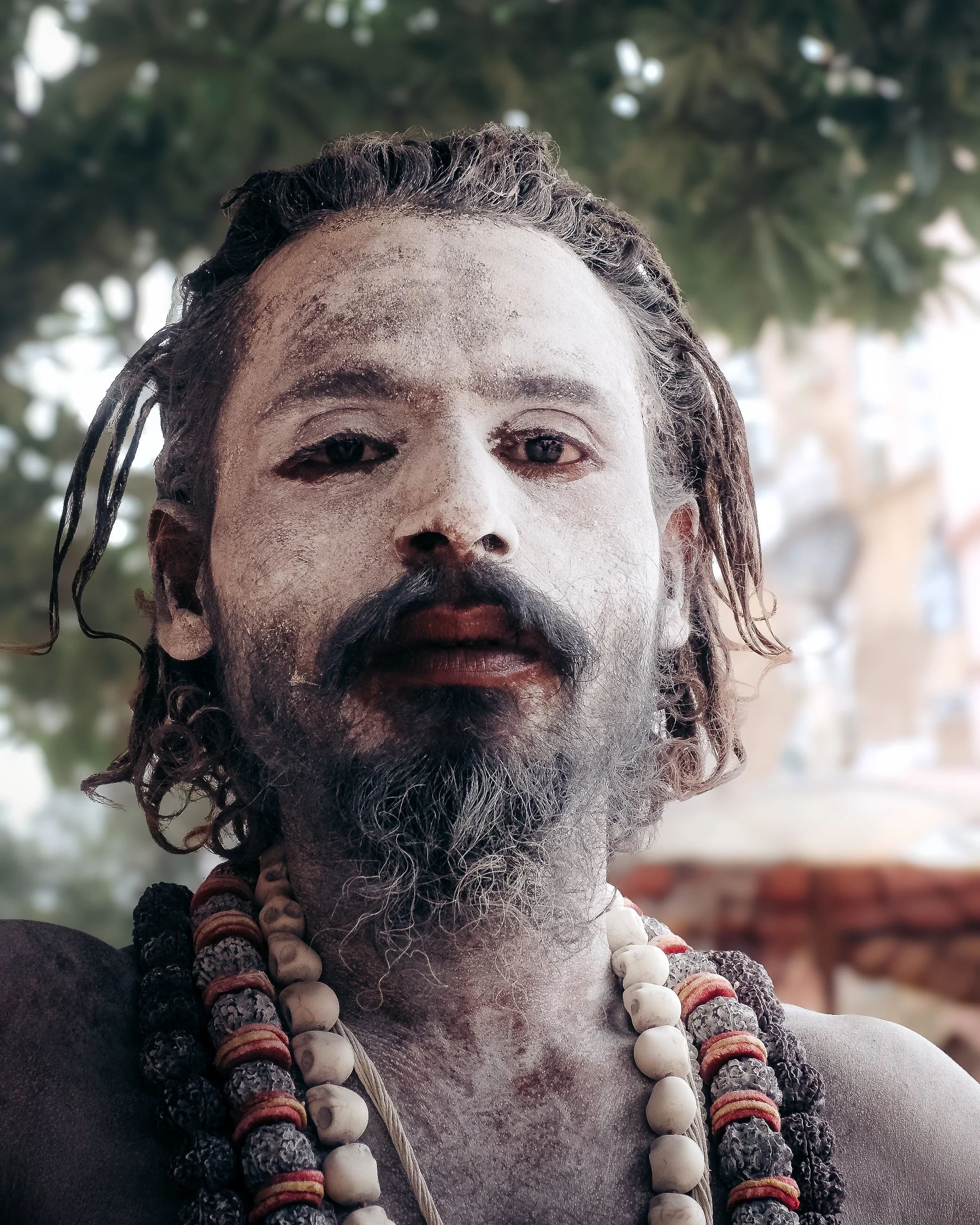 A person with ash-covered skin, long curly hair, and a beard, wearing multiple necklaces made of beads and skulls, standing outdoors with blurred greenery in the background.
