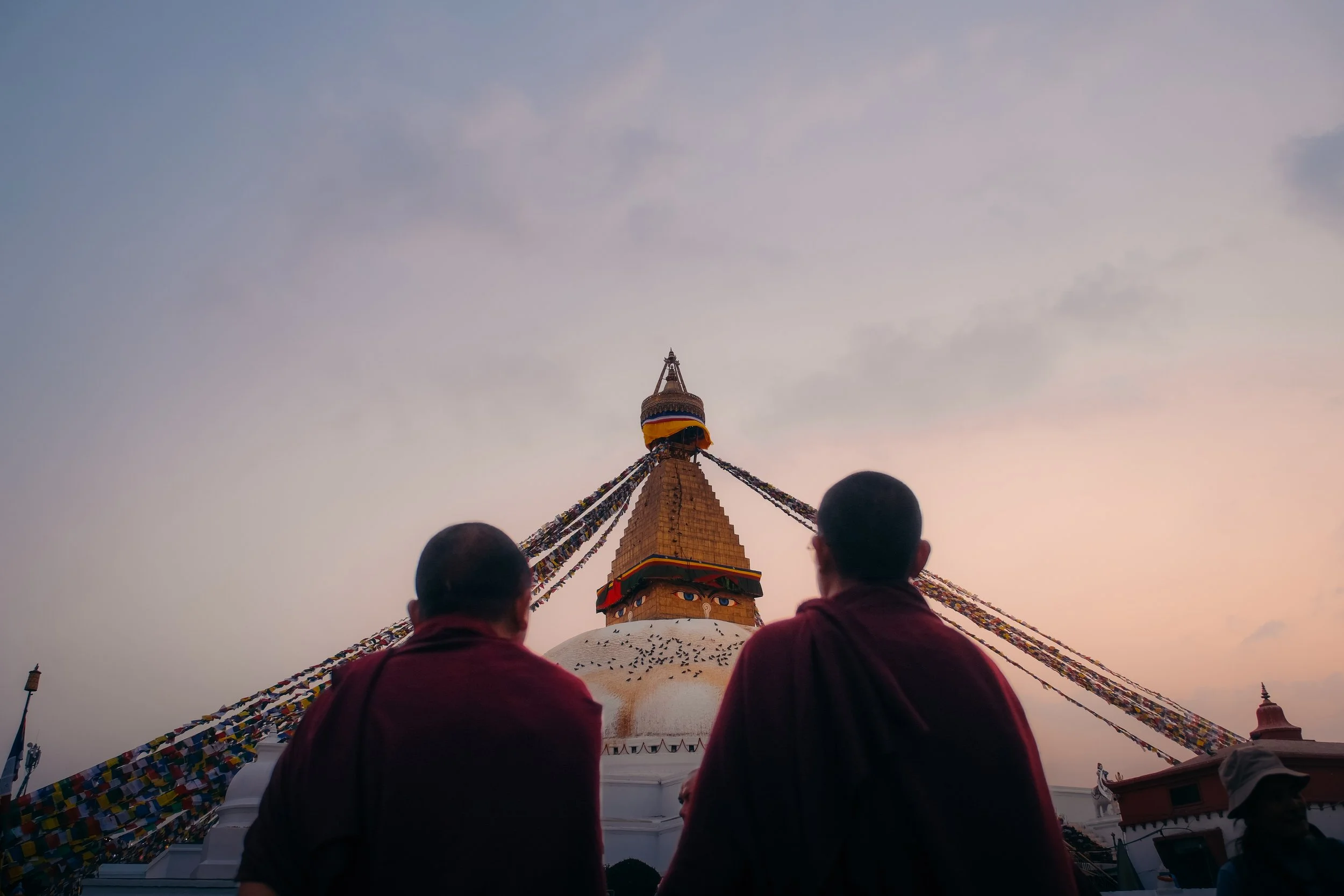 Two monks in maroon robes sit facing the Swayambhunath Stupa, also known as the Monkey Temple, in Kathmandu, Nepal, during sunset.