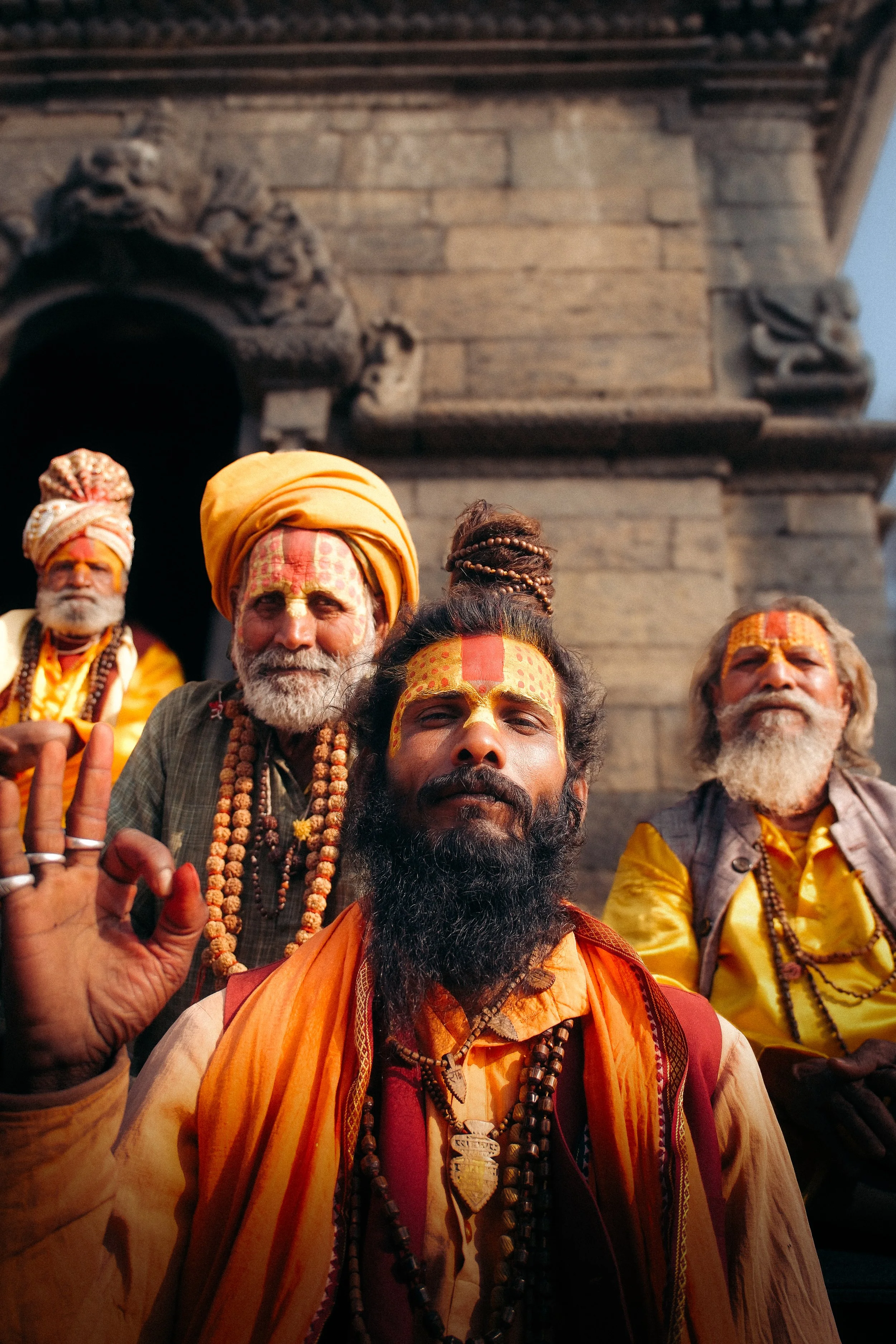 Four men with traditional Hindu attire and face paint, standing in front of a stone temple, one making an 'OK' gesture with his hand.