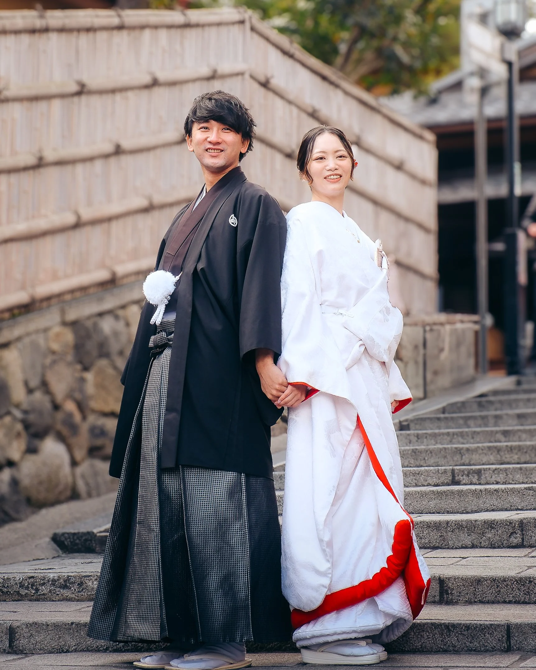 A couple in traditional Japanese wedding attire standing on stairs, holding hands, outdoors.