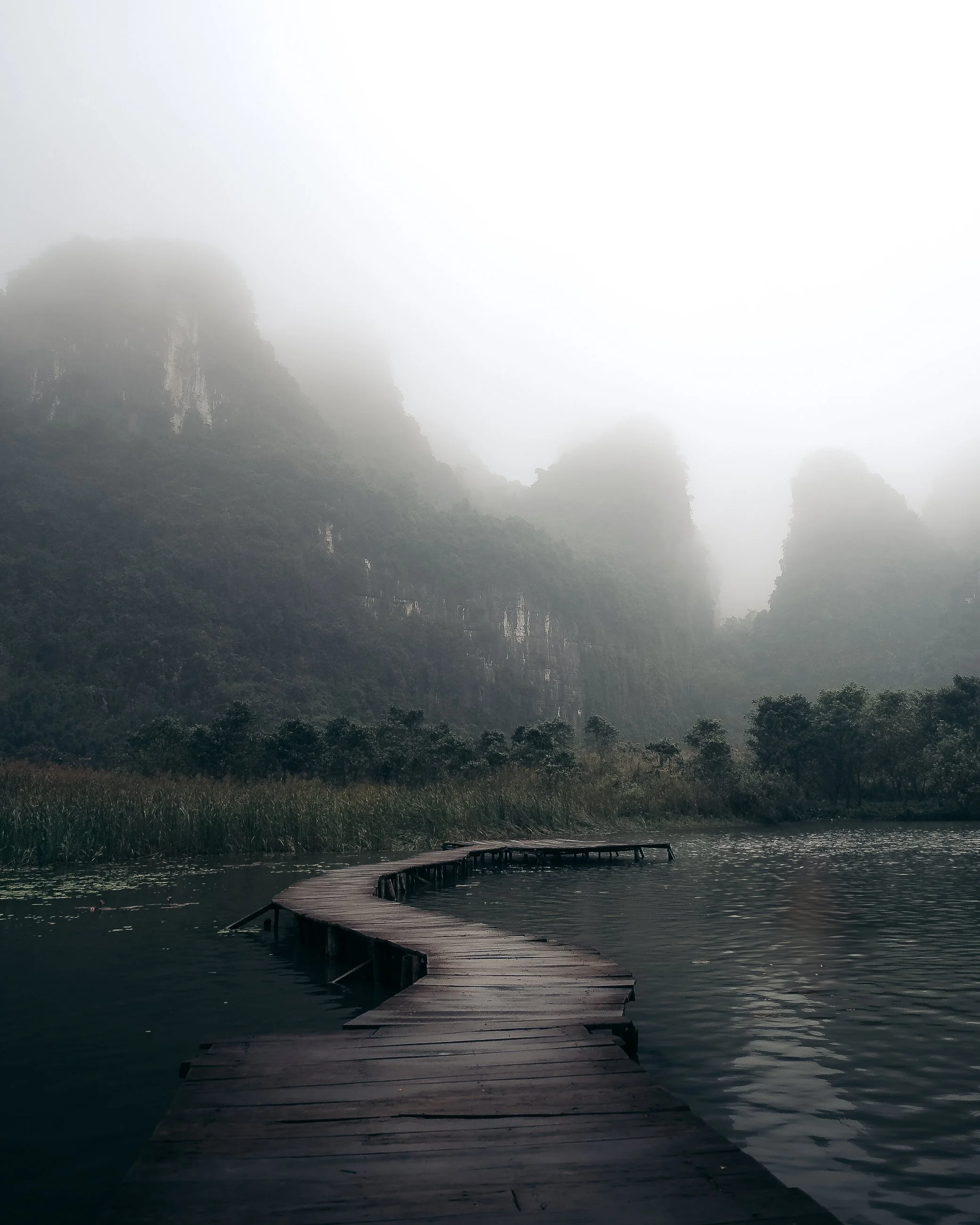 A winding wooden dock extends over a calm body of water, surrounded by foggy mountains and lush greenery.
