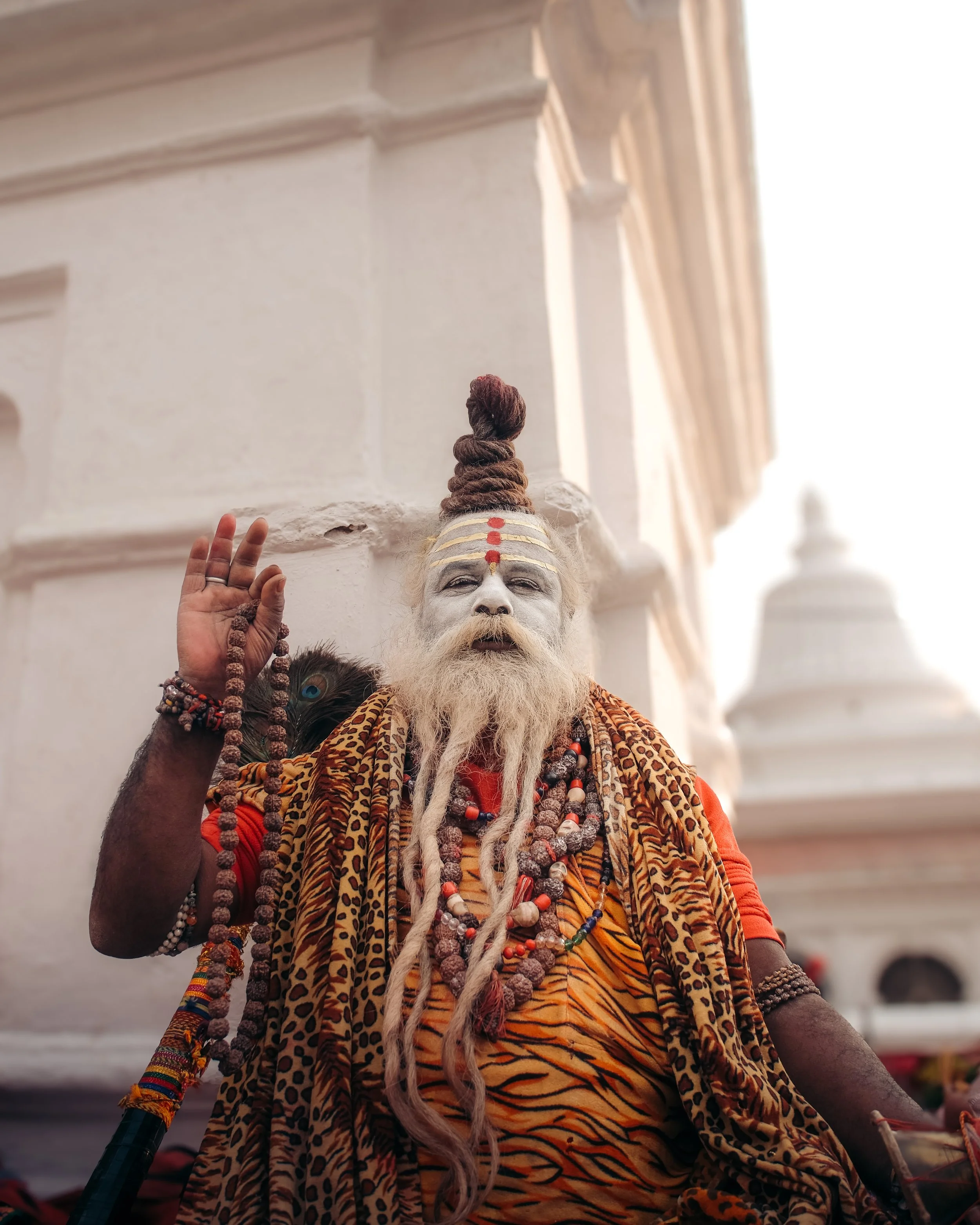 A person dressed as a Hindu spiritual figure with white face paint, long beard, and white hair styled in a topknot, wearing traditional robes with animal print, multiple necklaces, and holding prayer beads.