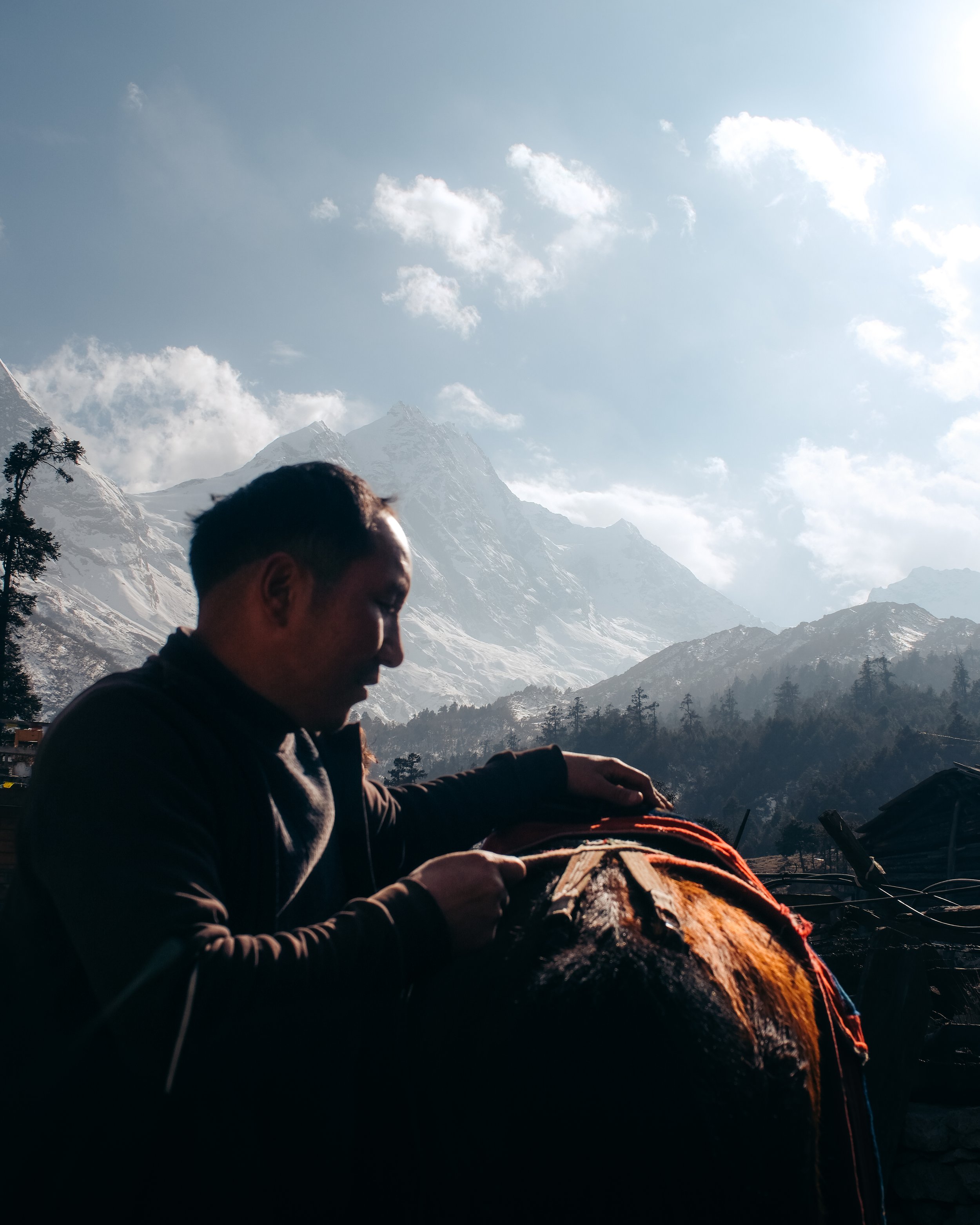 A man in a dark jacket stands outdoors in front of snow-capped mountains with a teepee or yurt in the background, holding a tool or object.