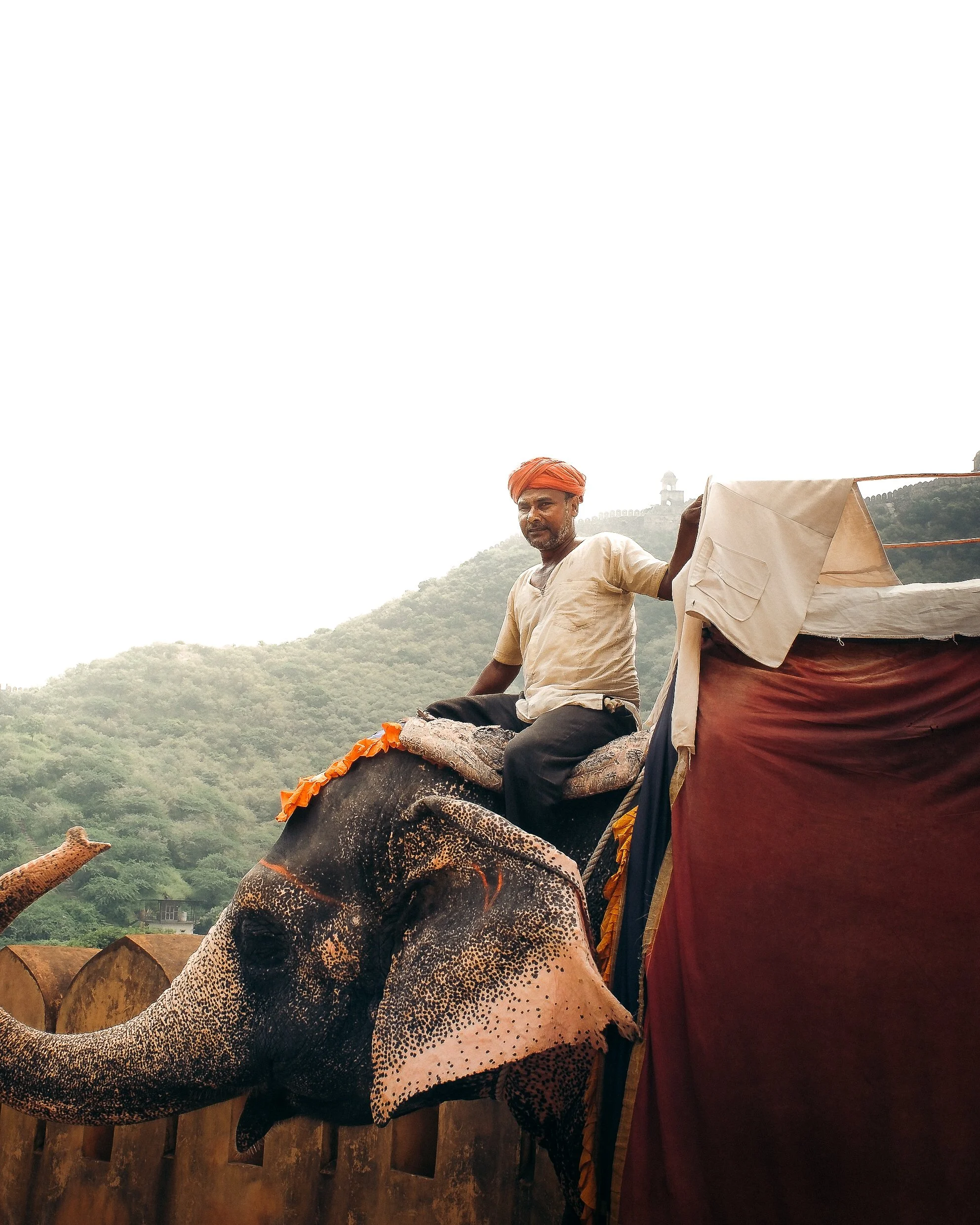 A man wearing a beige shirt and orange turban riding an elephant with decorated cloth, outdoors with hills and a building in the background.
