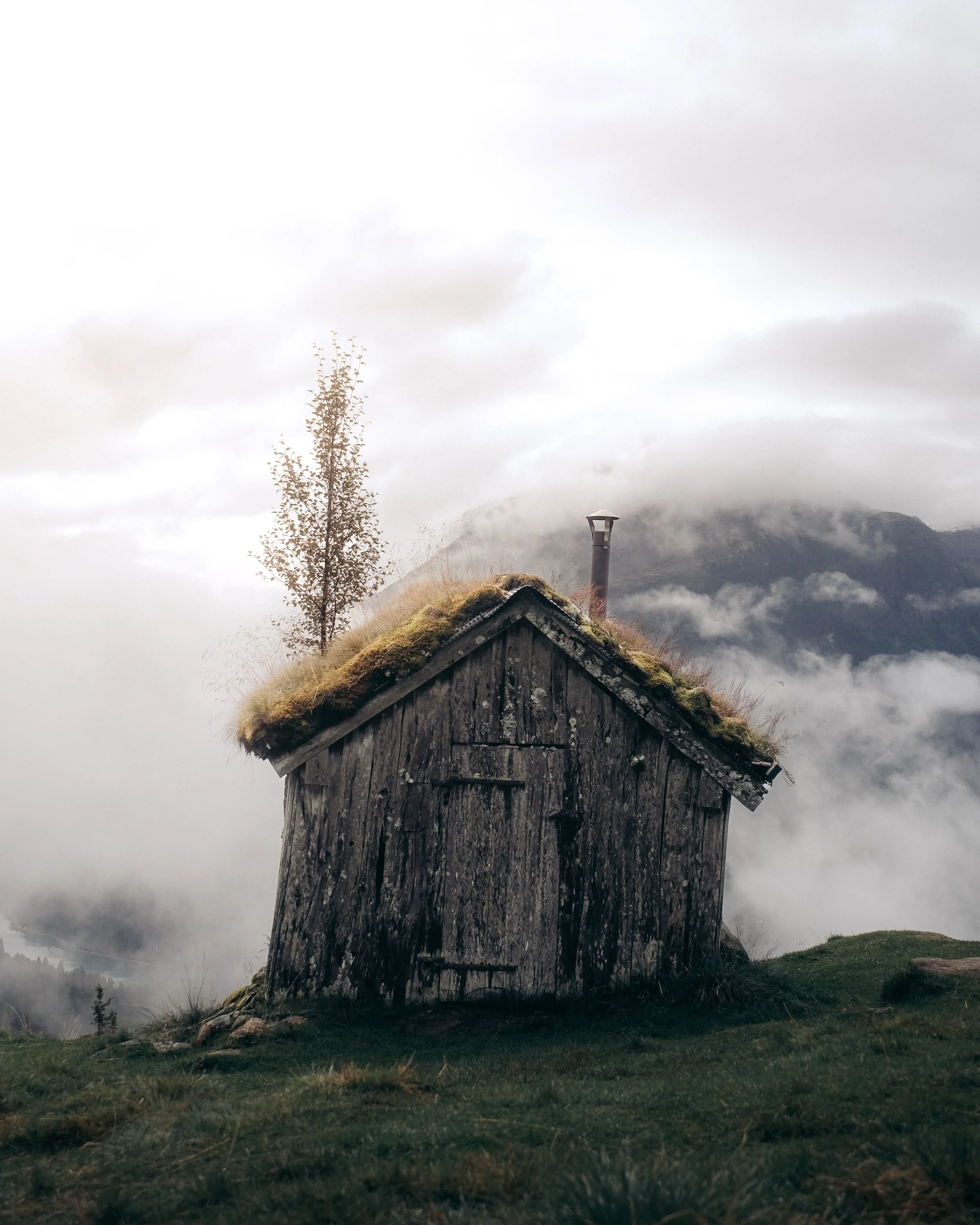 A small, weathered wooden hut with moss on the roof, set on grassy ground with a landscape of foggy mountains and cloudy sky in the background.