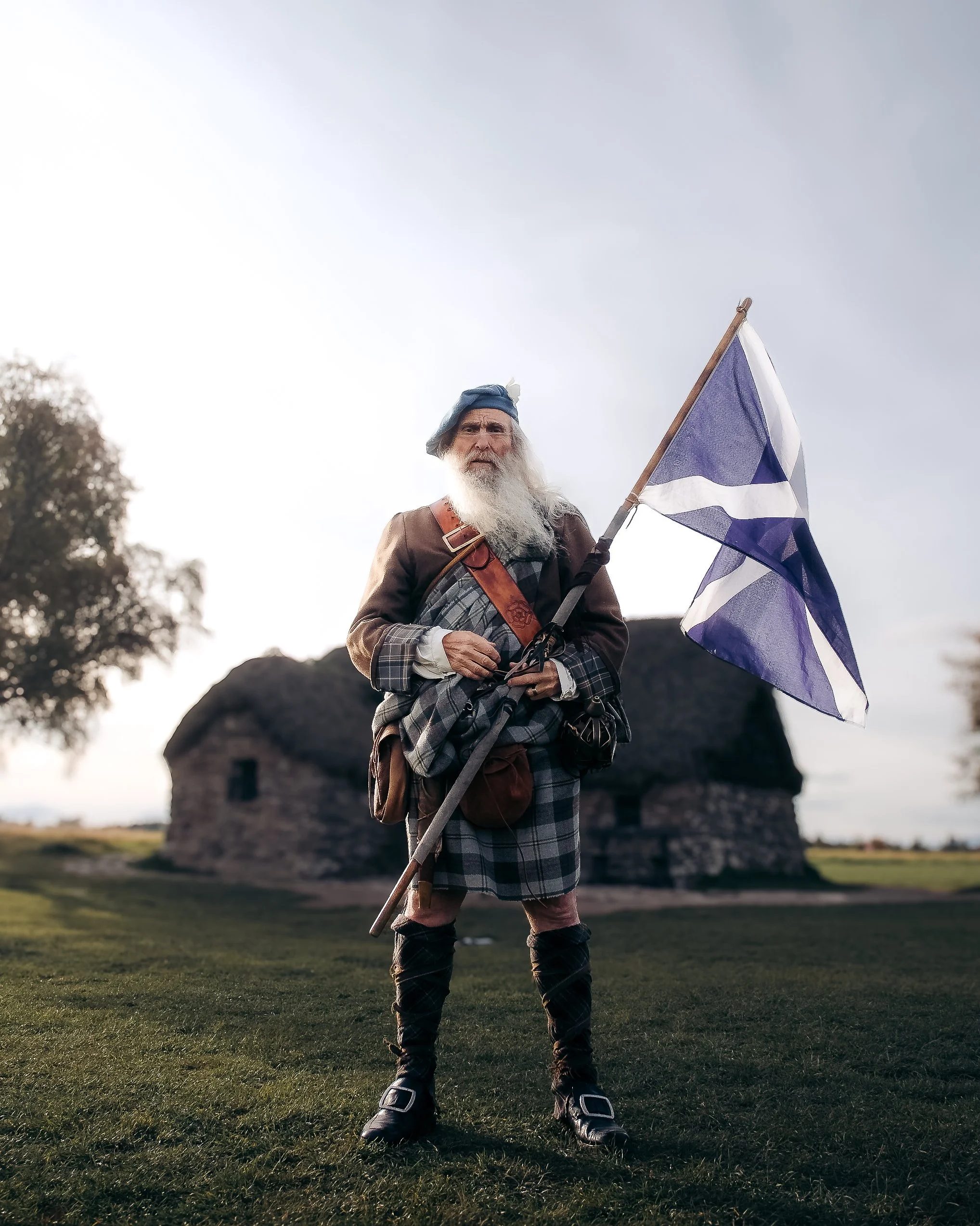 An elderly man dressed in Scottish traditional clothing standing outdoors on grass, holding a blue and white Scottish flag, with an old stone cottage and trees in the background during daytime.