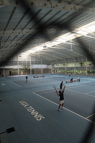 Indoor tennis court with multiple people playing and practicing. The court has the word 'SIMPLE' and 'LIFE' written on the surface. The ceiling is high with metal beams and natural light coming through windows.
