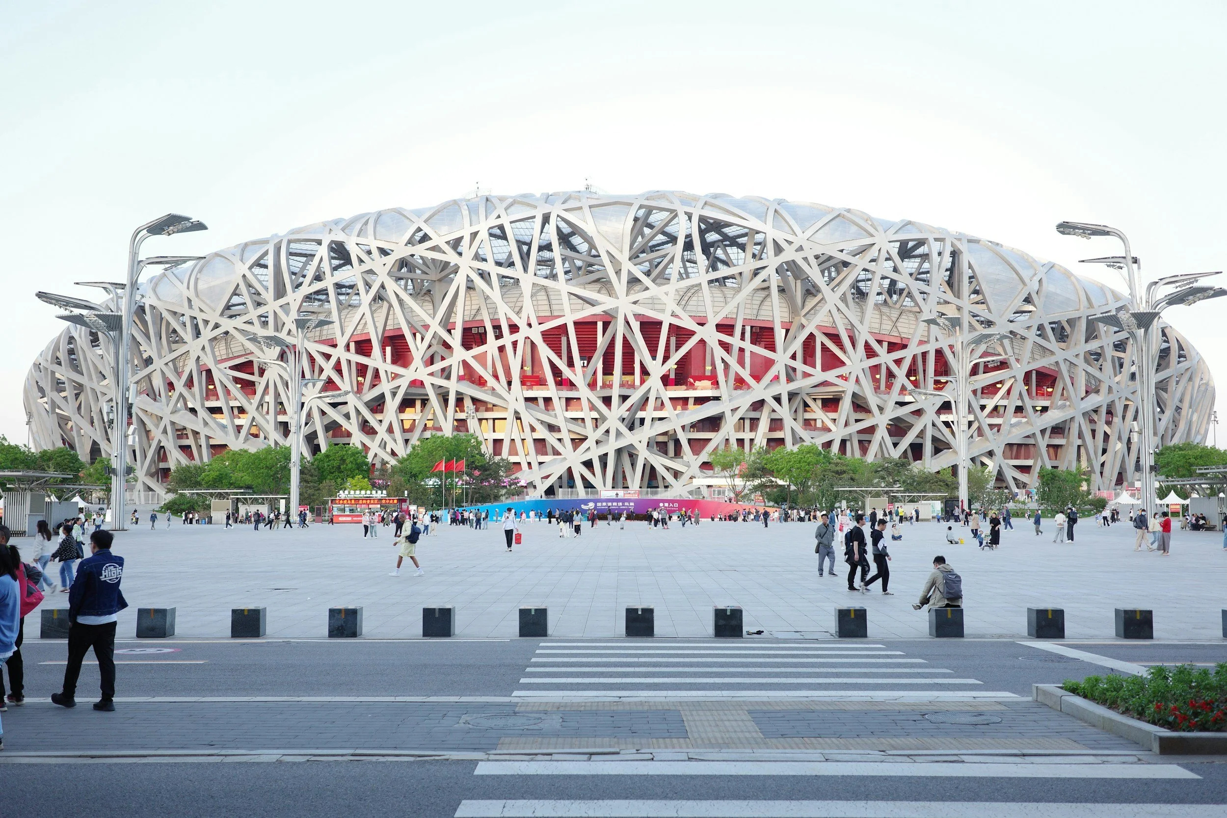 Modern stadium with an intricate lattice exterior, surrounded by people and street lamps, under a pale sky.