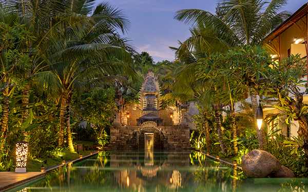A tropical resort pool area with lush palm trees, stone steps, and a small waterfall, illuminated by warm lighting at dusk.