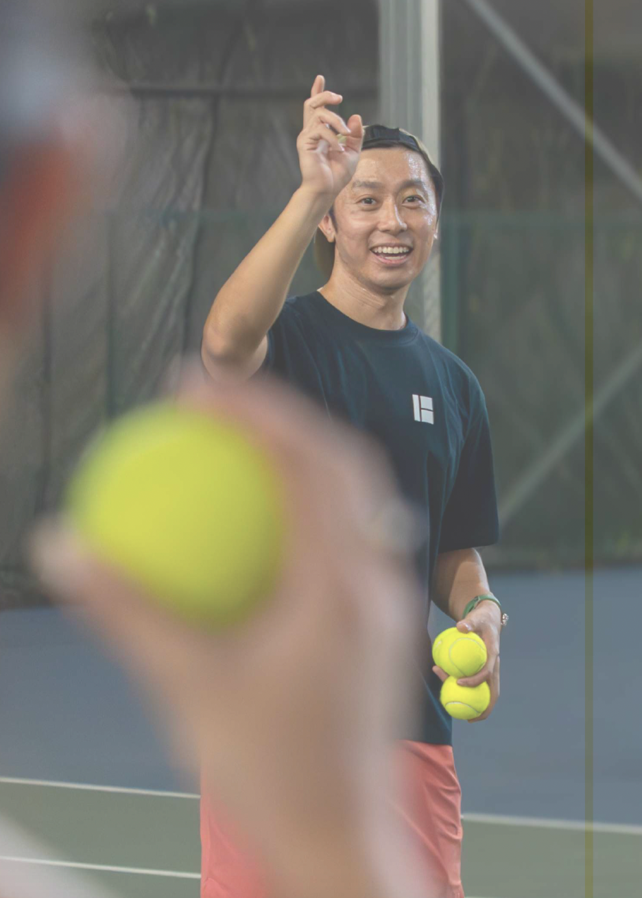 A man holding tennis balls on a tennis court, smiling, with another person's blurred arm and tennis ball in the foreground.