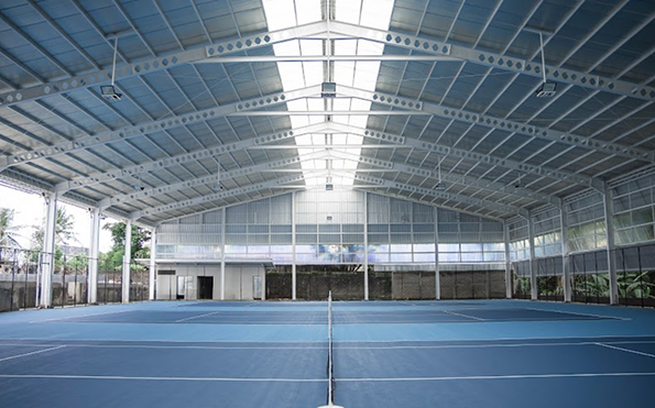 Indoor tennis court with blue playing surface under a high, metal, arched roof.