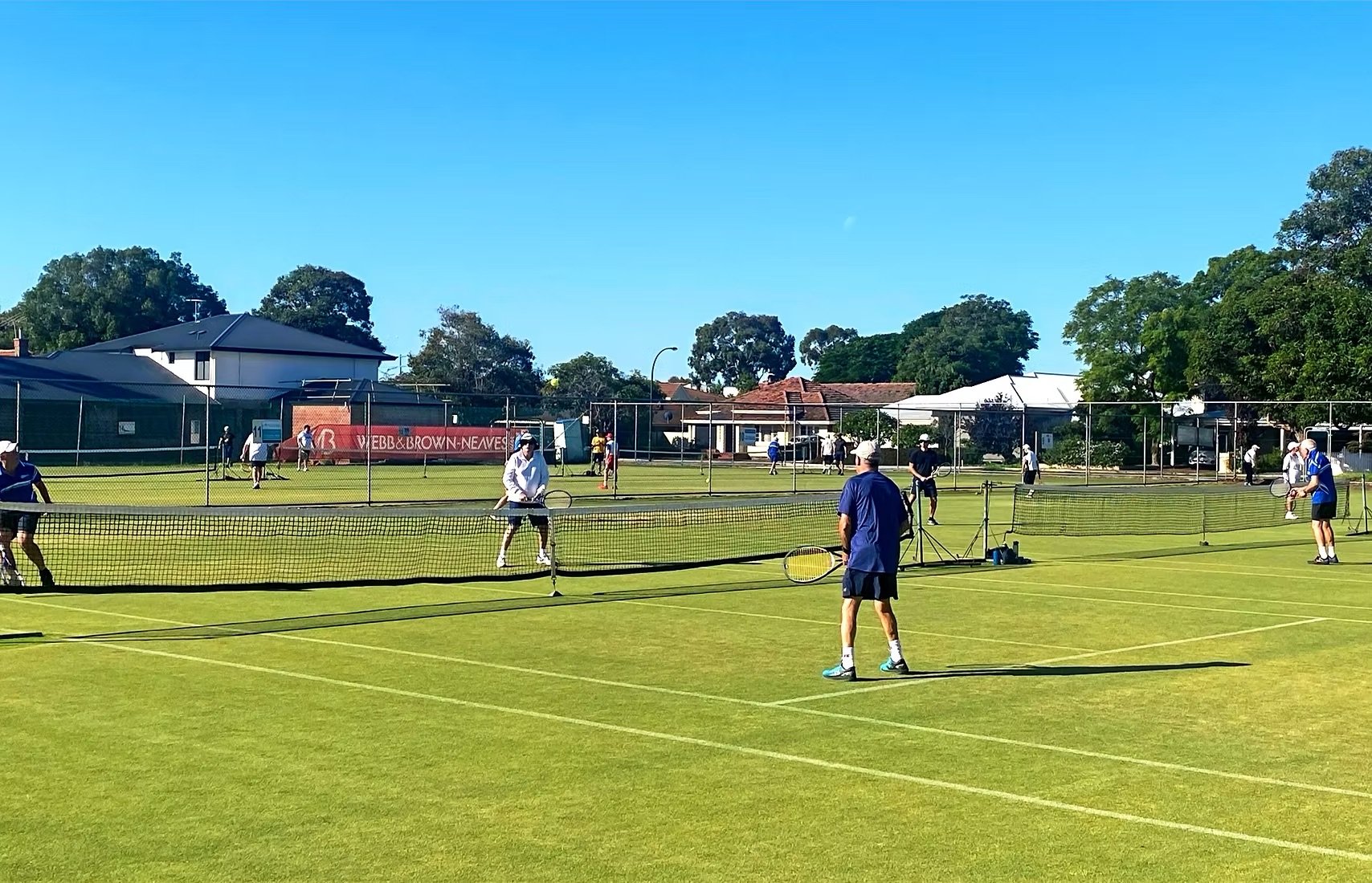 People playing tennis on multiple courts in a park on a sunny day with trees and houses in the background.