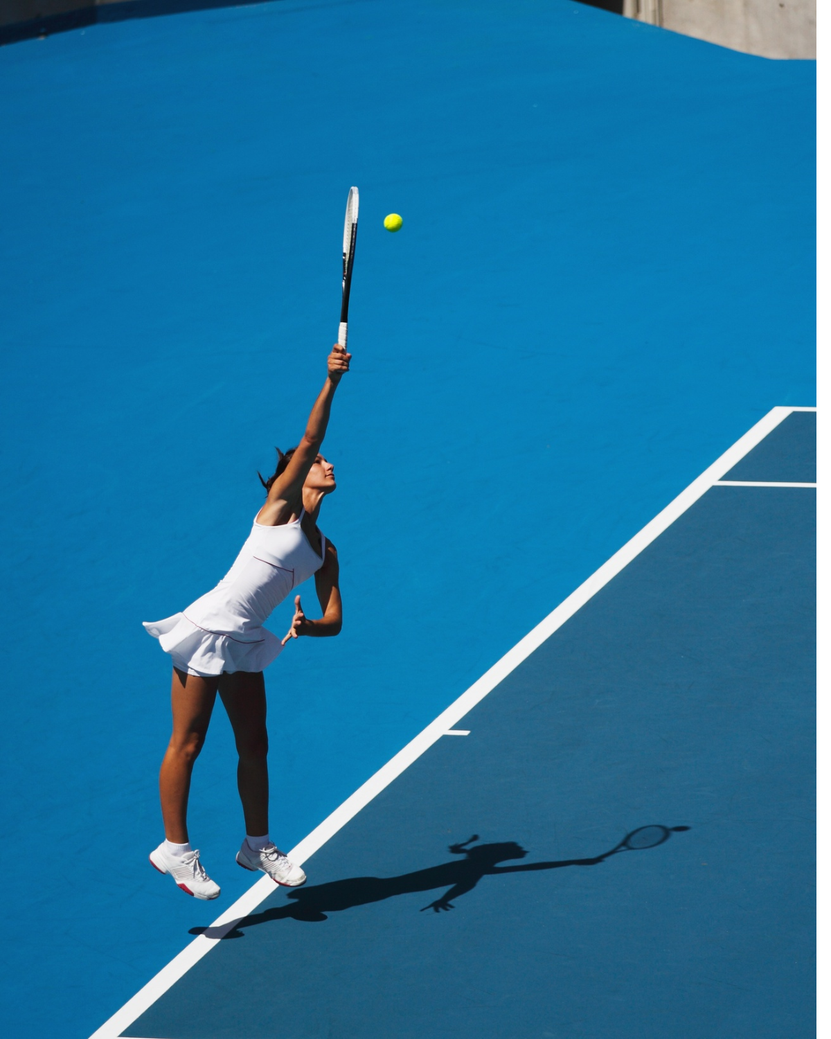 A female tennis player in a white tennis outfit hitting a tennis ball on a blue tennis court