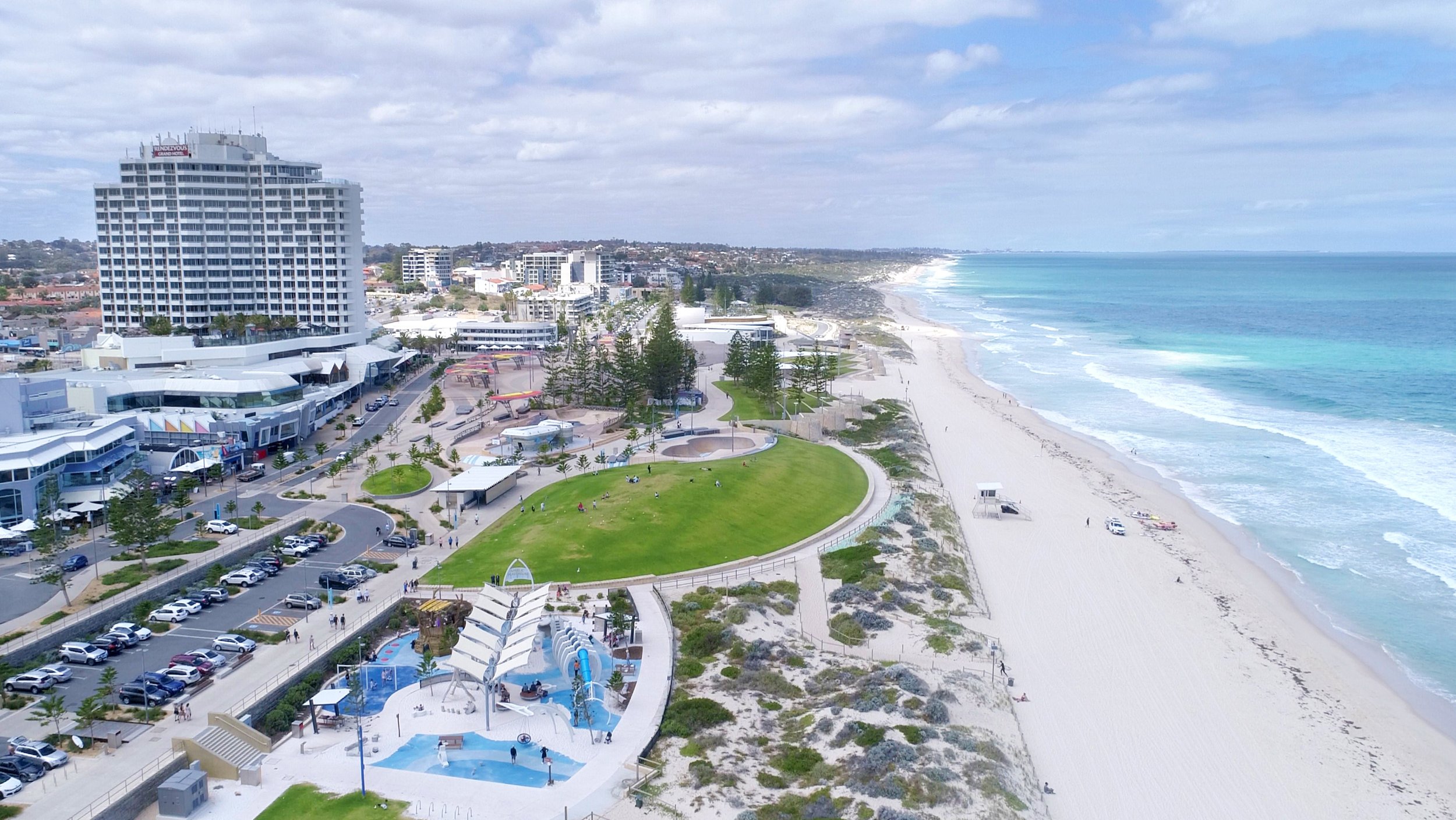 A coastal cityscape with a beach, high-rise buildings, a park, and a water park along the shoreline under a partly cloudy sky.