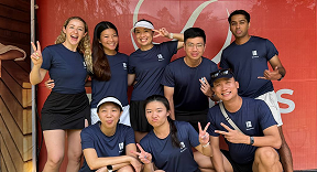 Group of nine young people in matching navy blue t-shirts and black shorts posing together indoors in front of a red wall with a partial logo, some making peace signs.