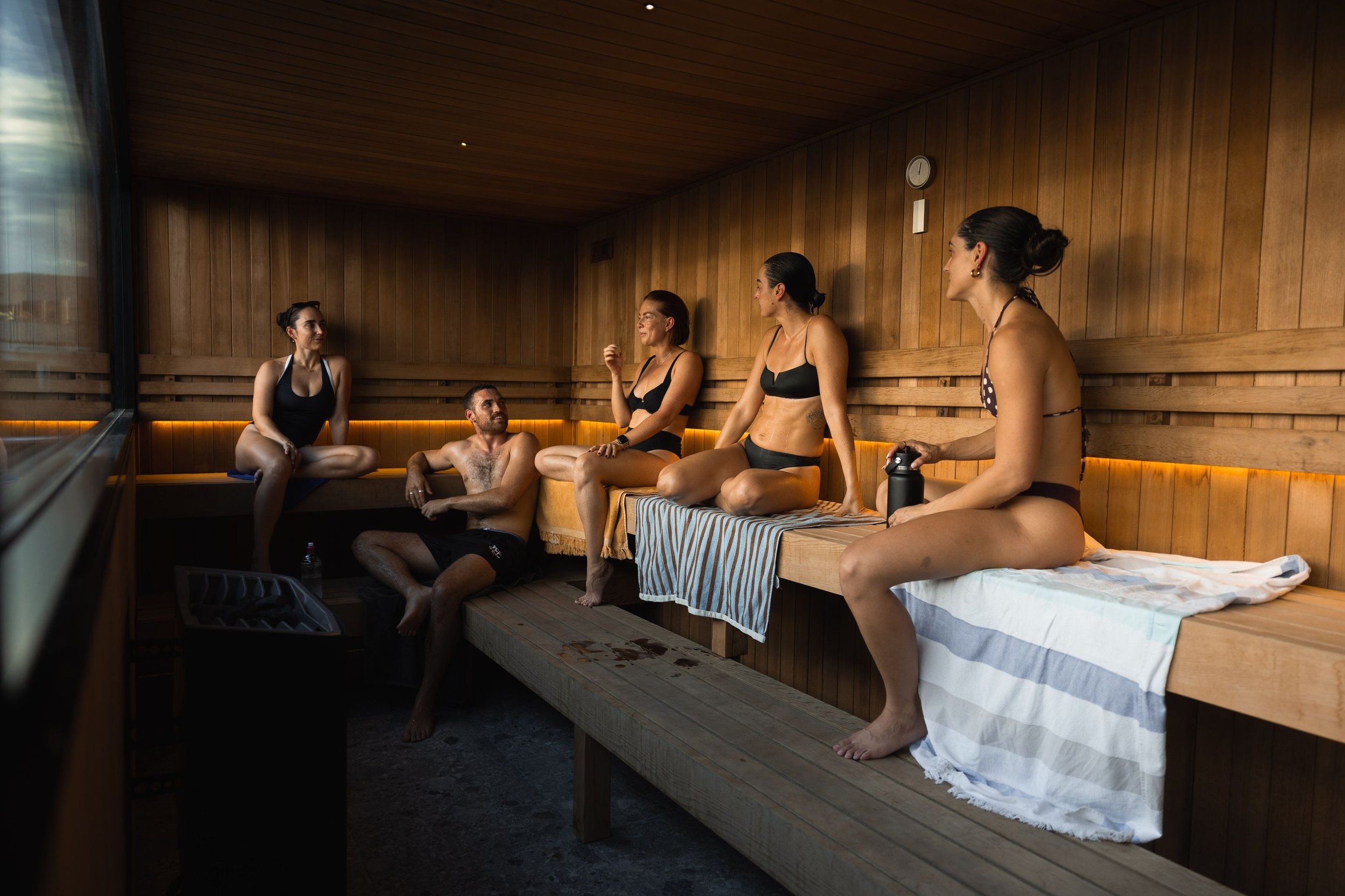 Six people relaxing in a traditional wooden sauna, with five women dressed in swimsuits and one man sitting on the floor, engaging in conversation.