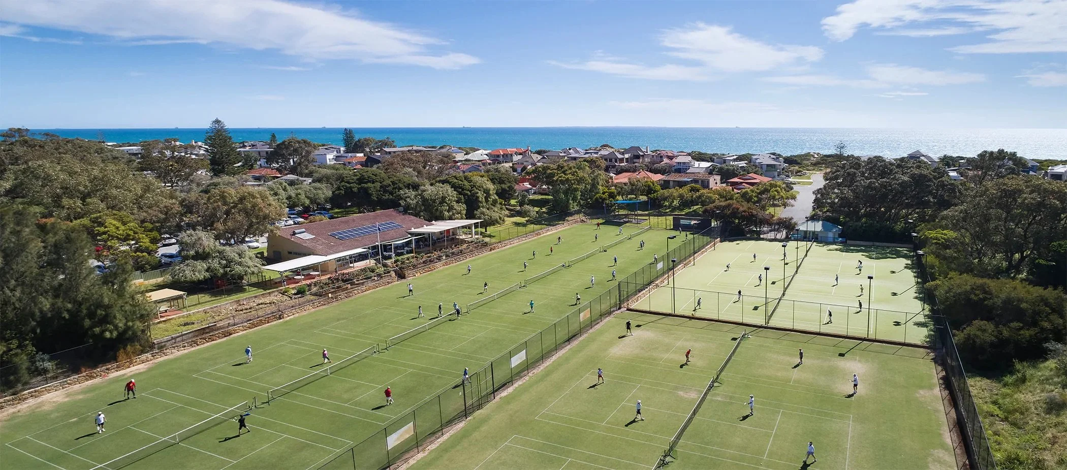 An aerial view of a tennis facility with multiple courts near the coast. Several people are playing tennis, and there is greenery and residential houses in the background with the ocean and blue sky beyond.