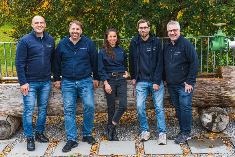 Fünf Personen stehen nebeneinander vor einer Baumstamm, umgeben von Herbstblättern, alle tragen dunkle Jacken mit Firmenlogo der Reatech AG. Reatech AG ist die Firma hinter dem Floating Tank Rotkreuz (Rea Float)