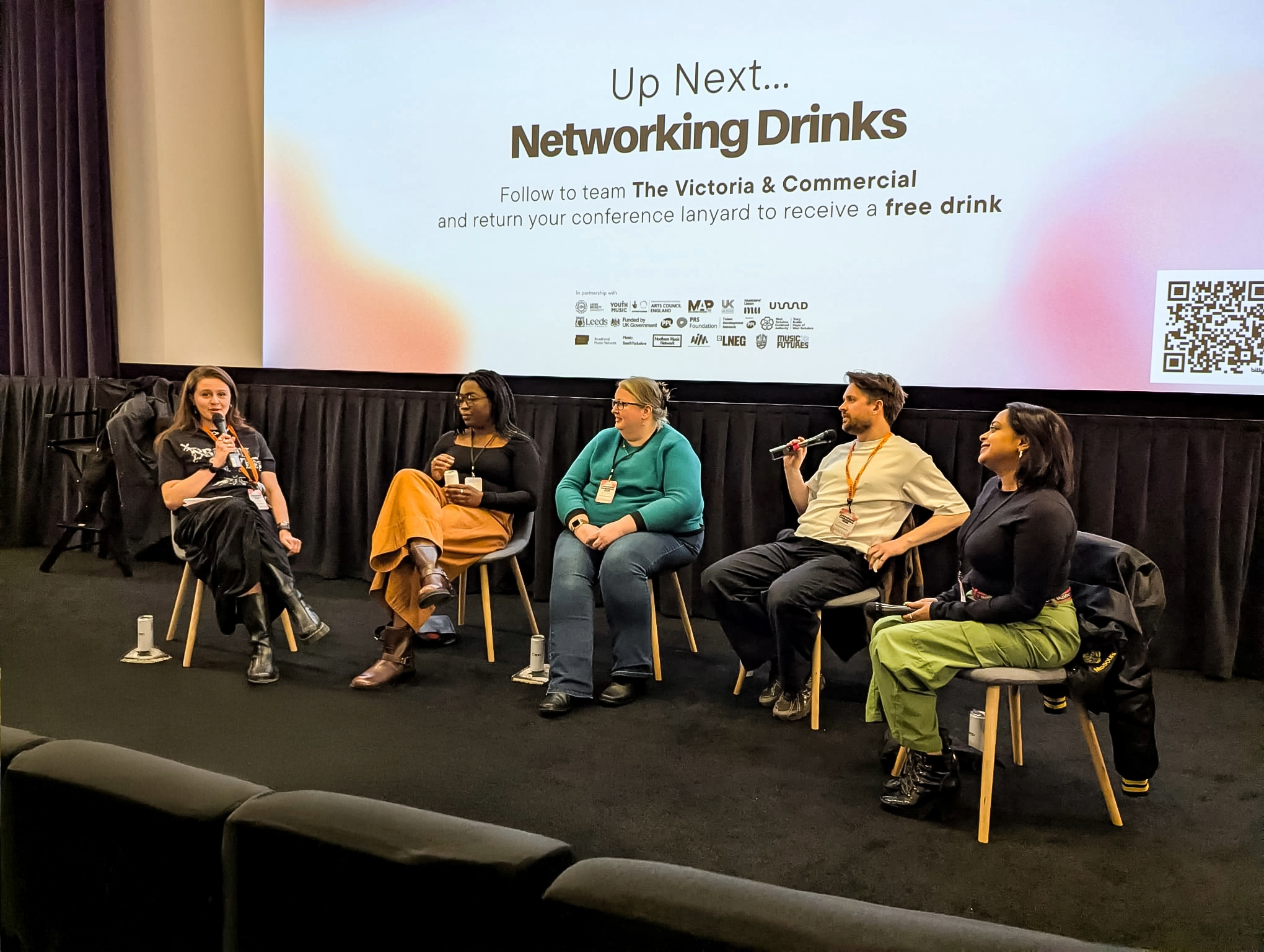Group of five individuals seated on stage in front of a large presentation screen, engaging in panel discussion. The screen displays a message about networking drinks to follow.