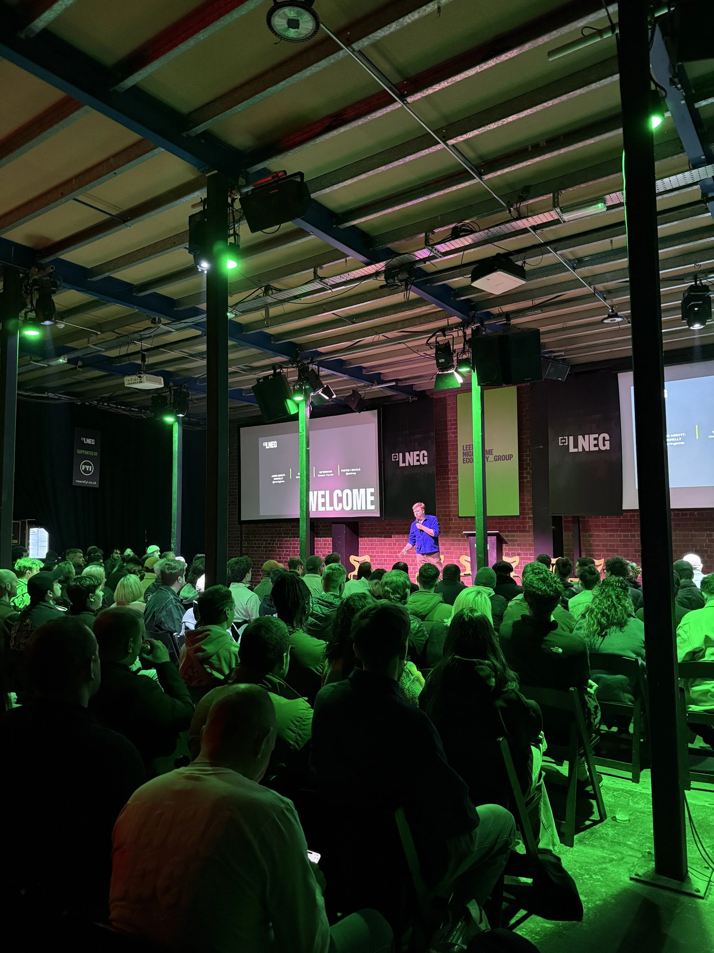 An audience attending a conference or seminar in a dimly lit room with green stage lighting. A speaker stands at a podium in front of a brick wall, with multiple screens displaying logos and the word 'WELCOME - LNEG'.