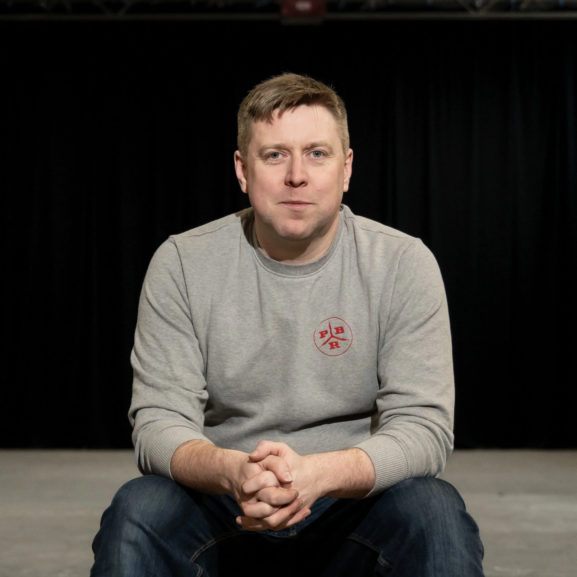 A man with short light brown hair and fair skin, wearing a gray sweatshirt with red circular logo on the chest, sitting with his hands clasped in front of him, in front of a black curtain background.