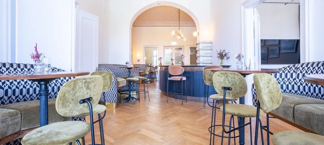 Innenraum eines Cafés mit Tischen, Stühlen und Blumen, helles Dekor, Holzboden, gebogene Bar mit Stühlen im Hintergrund.