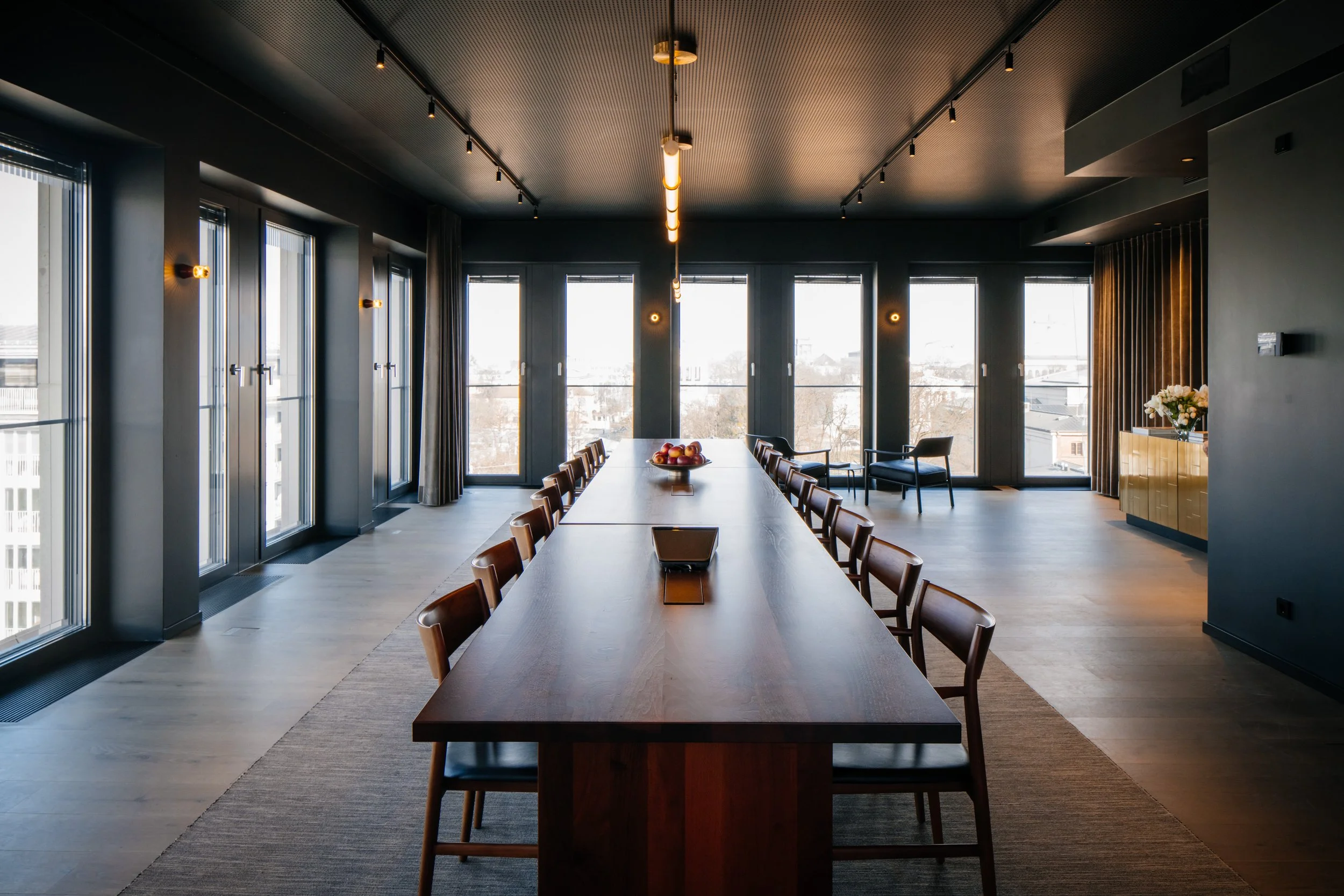 Ein moderner, heller Konferenzraum mit großem langen Tisch, Stühlen, großen Fenstern, einer Deckenlampe und einer Sideboard mit Blumen