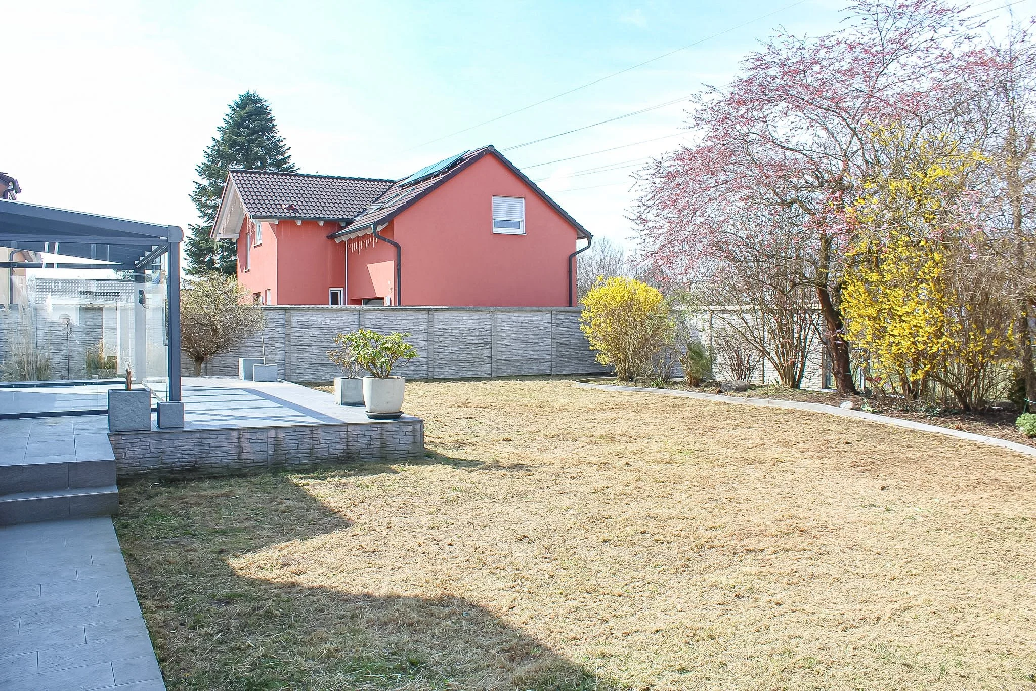 Ein Garten mit einem Rasen, mehreren Bäumen, einem modernen Haus mit rot-orangefarbenen Wänden und einem Dach mit Solarpanelen, sowie einem gemauerten Zaun und einem teilsüberdachten Sitzbereich.