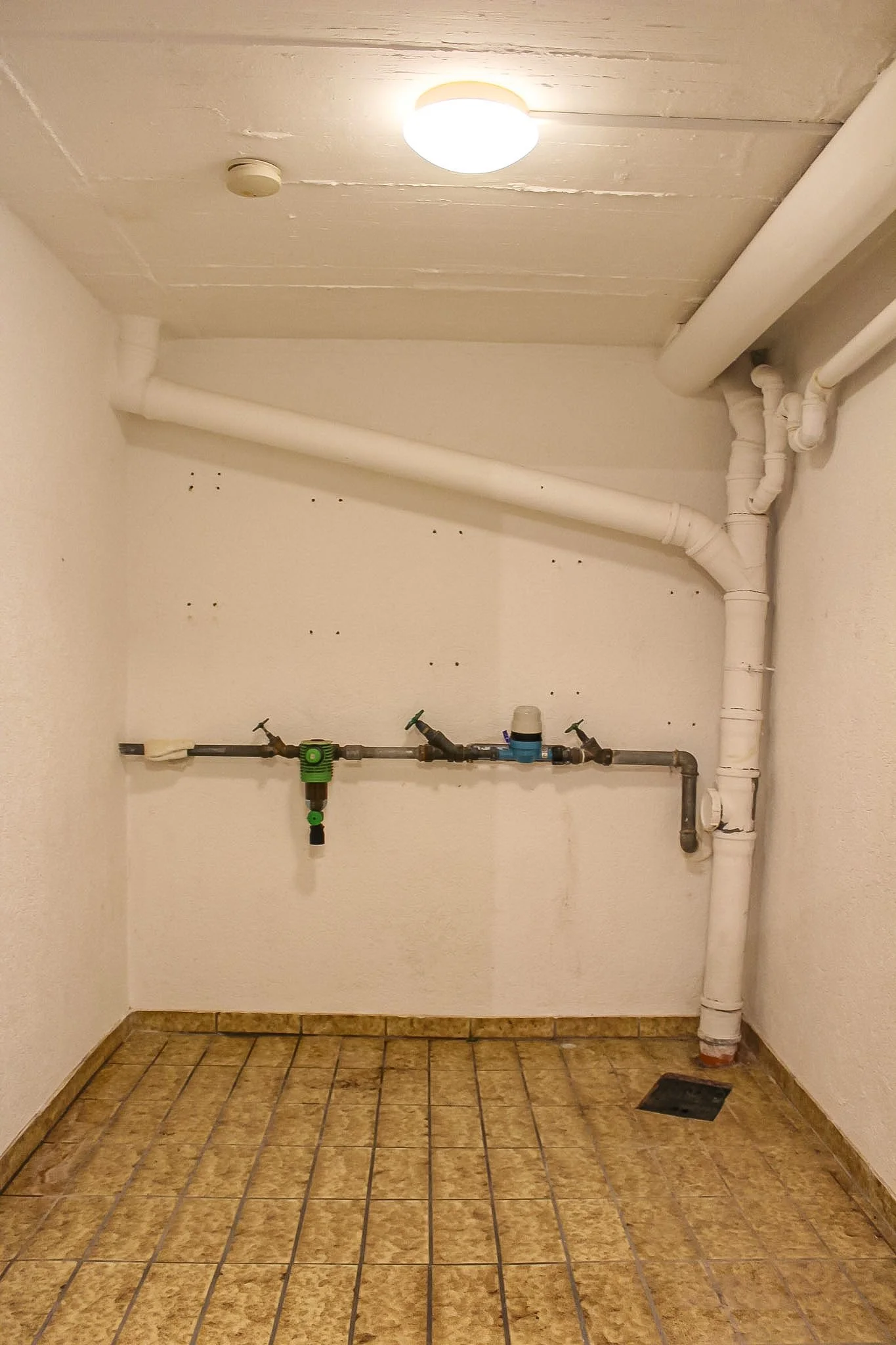 Innenaufnahme eines Raumes mit weißen Wasser- und Abflussrohren, einer Kontrollbox und einem kleinen Ablauf im Boden, die Wände sind weiß, der Boden ist gefliest.