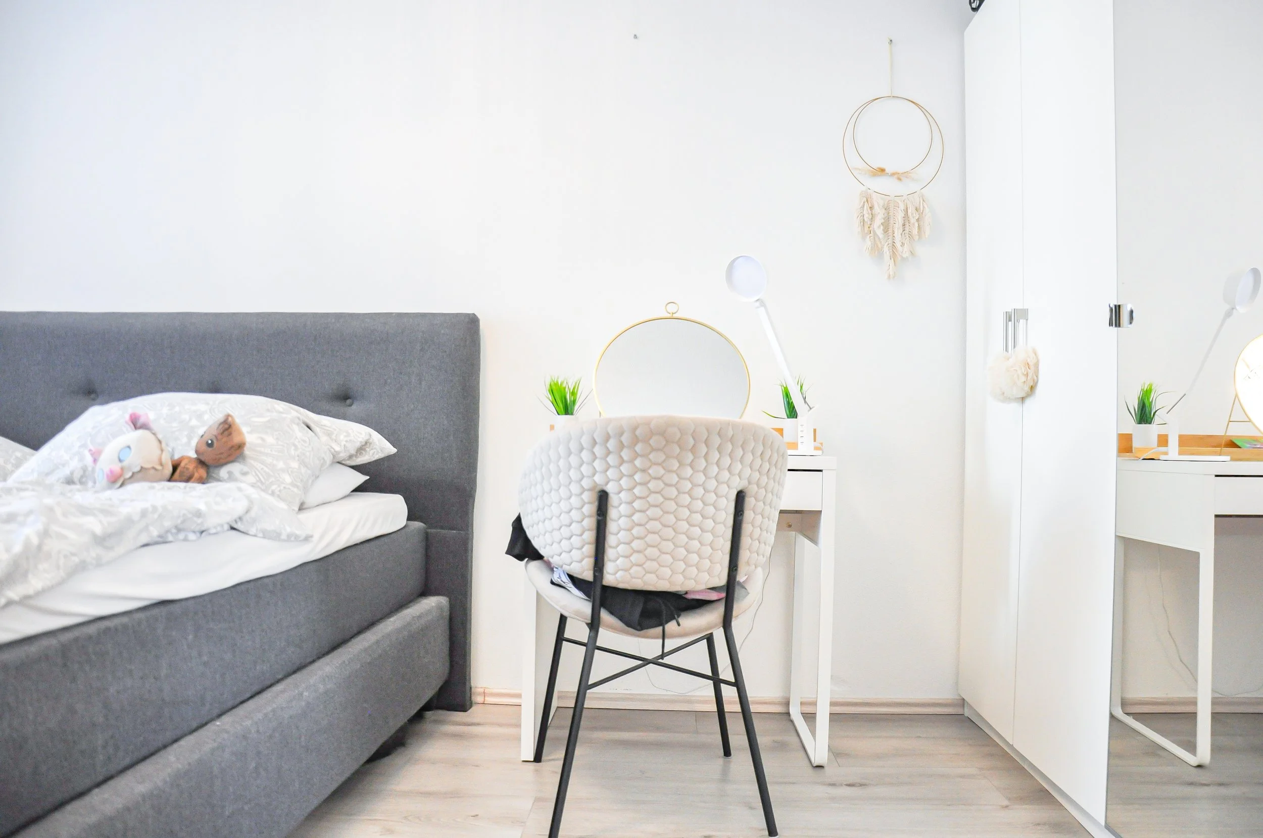 Schlafzimmer mit grauem Bett, weißen Bettwäsche, Kuscheltieren, weißen Wanddekorationen, Schreibtisch mit Pflanzen, Spiegel, Beleuchtung.