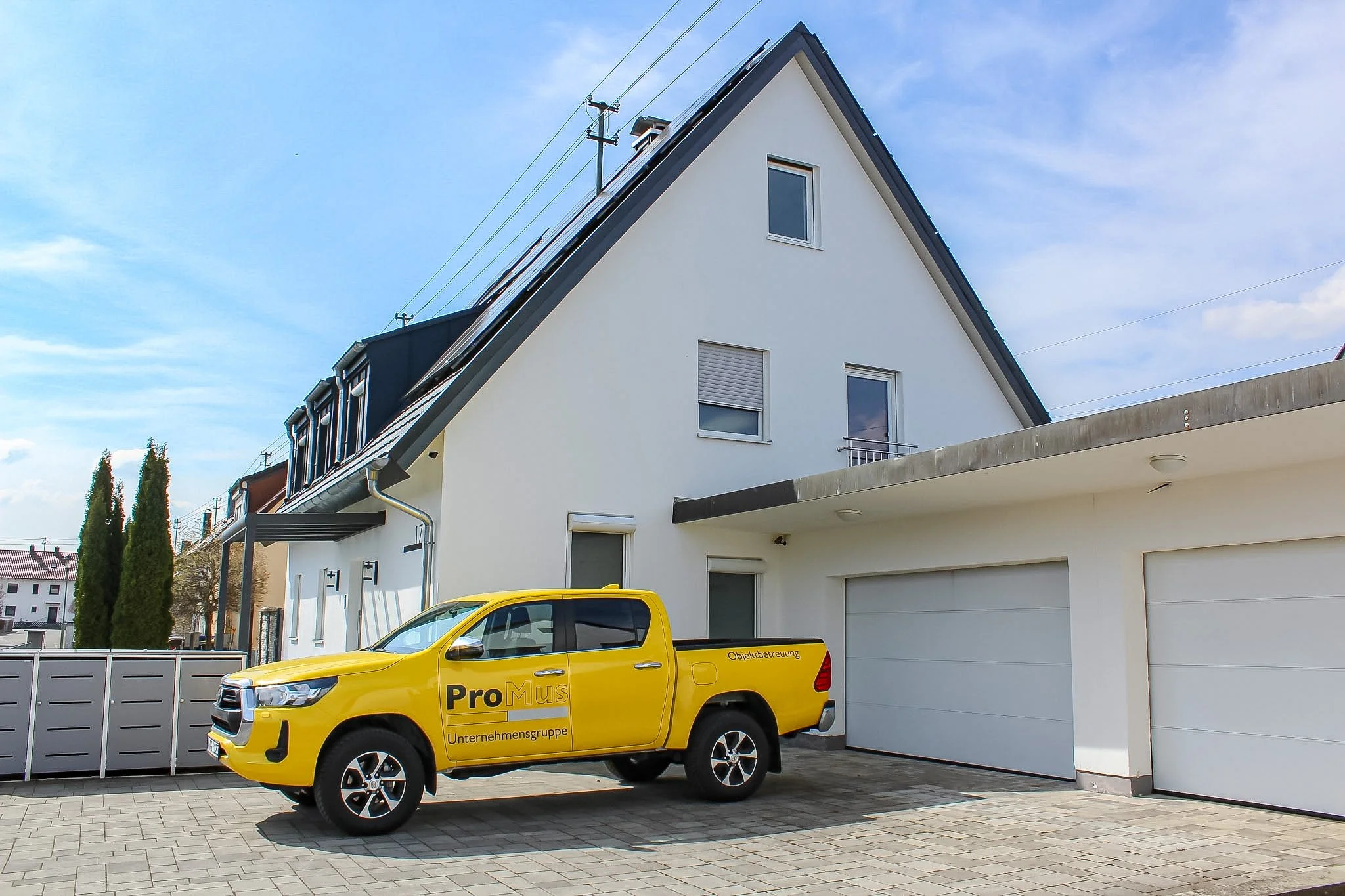 Gelber Unternehmenswagen mit Aufschrift "Pro Mus" vor einem modernen weißen Haus mit Satteldach und mehreren Fenstern bei Sonnenwetter.