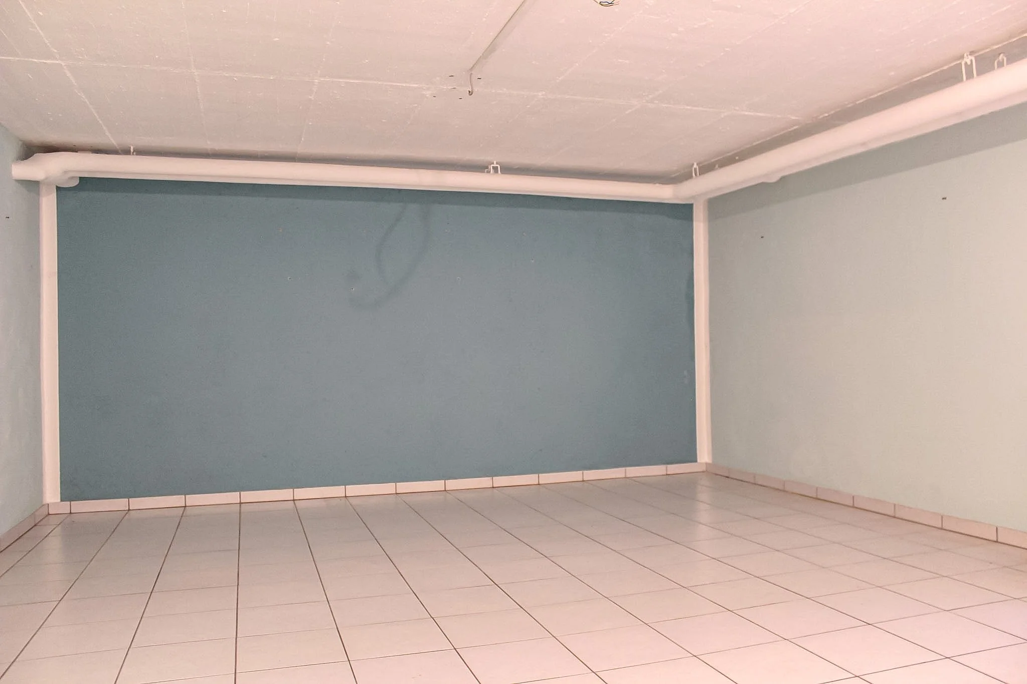 Ein leerer Raum mit blauer Wand, weißen Fliesen und sichtbarer Decke mit Rohrleitungen.