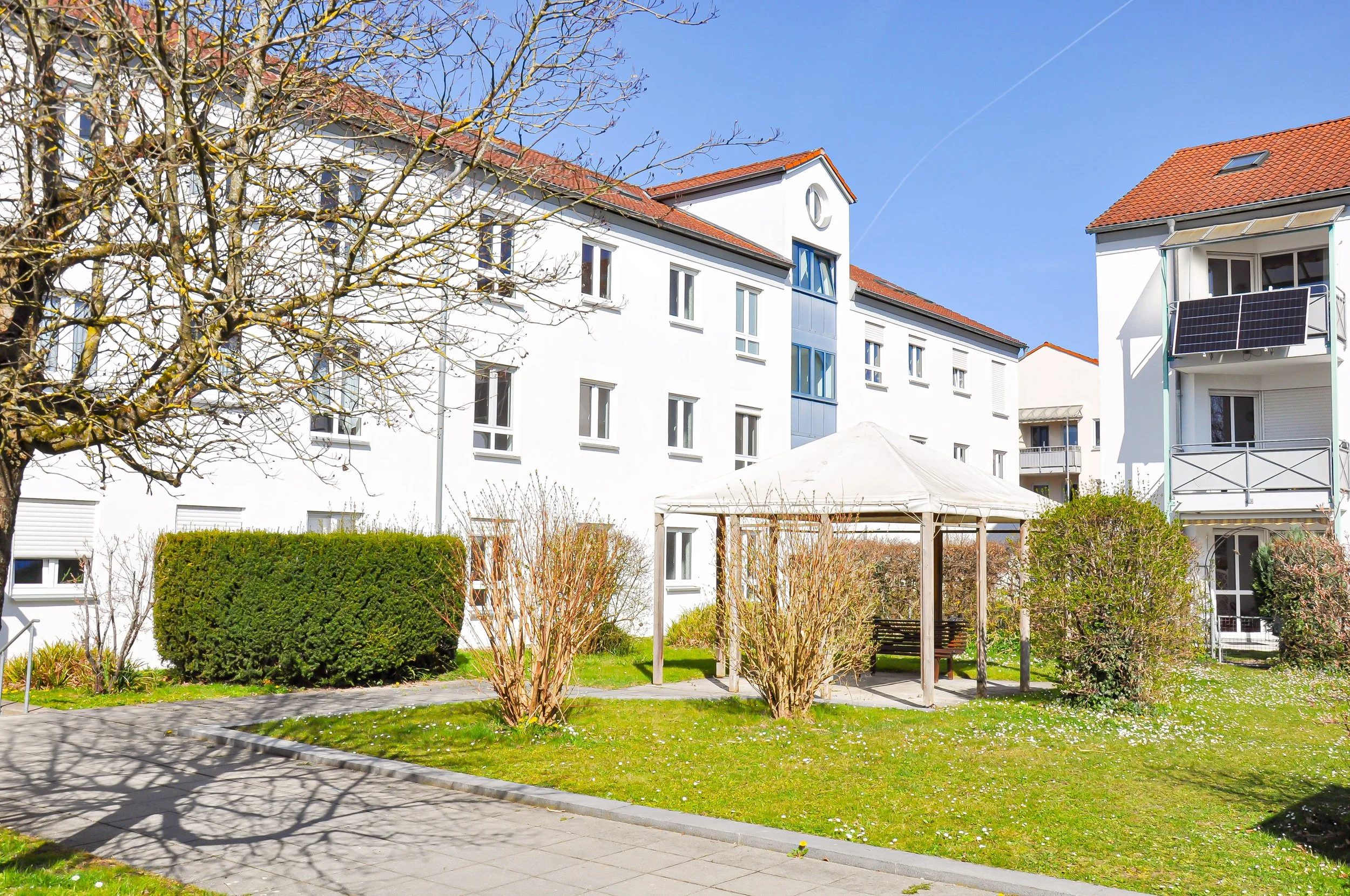 Mehrfamilienhaus mit roten Ziegeldächern, Solarzellen, und einem Garten mit Bäumen und einem Pavillon bei schönem Wetter.