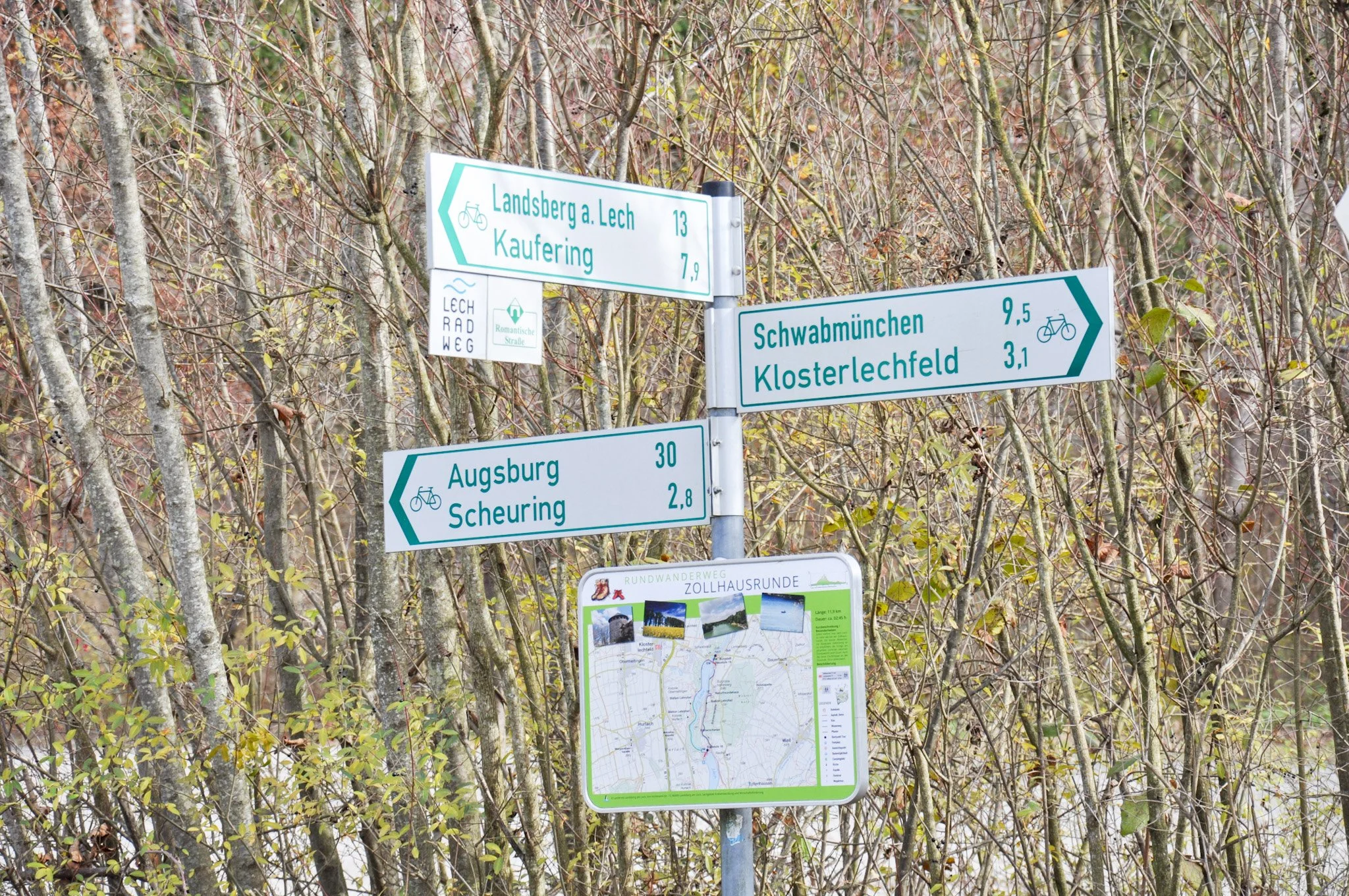 Mehrere Wegweiser mit Radwegnamen und Entfernungen stehen vor einem Baumstrauch. Die Wegweiser zeigen nach Landsberg, Kaufering, Schwabmünchen, Klosterlechfeld, Augsburg und Scheuring.