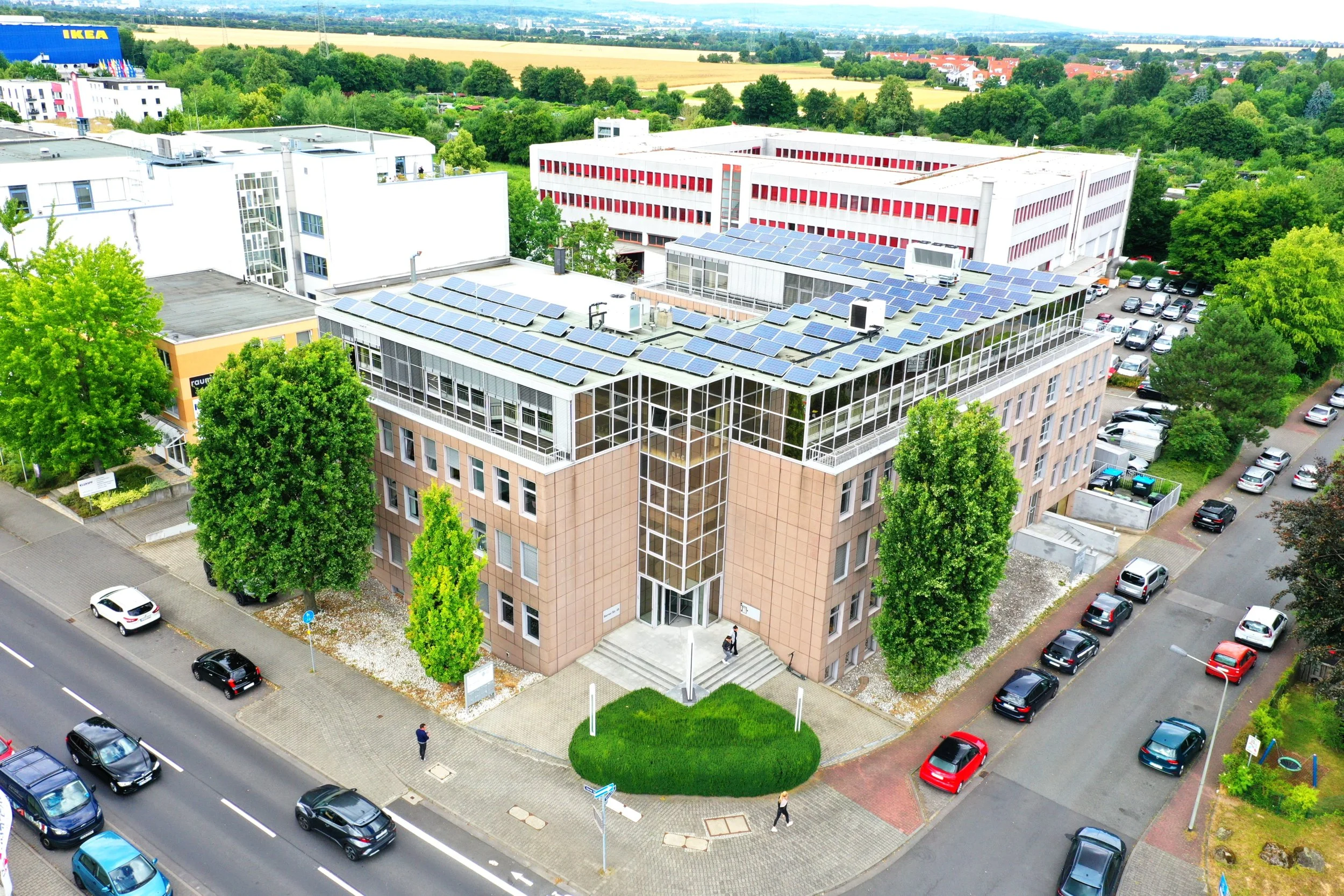 Luftaufnahme eines modernen Bürogebäudes mit Solarpaneelen auf dem Dach, umgeben von Parkplatz und grünen Bäumen in einem urbanen Bereich, im Hintergrund landwirtschaftliche Flächen und Bäume.