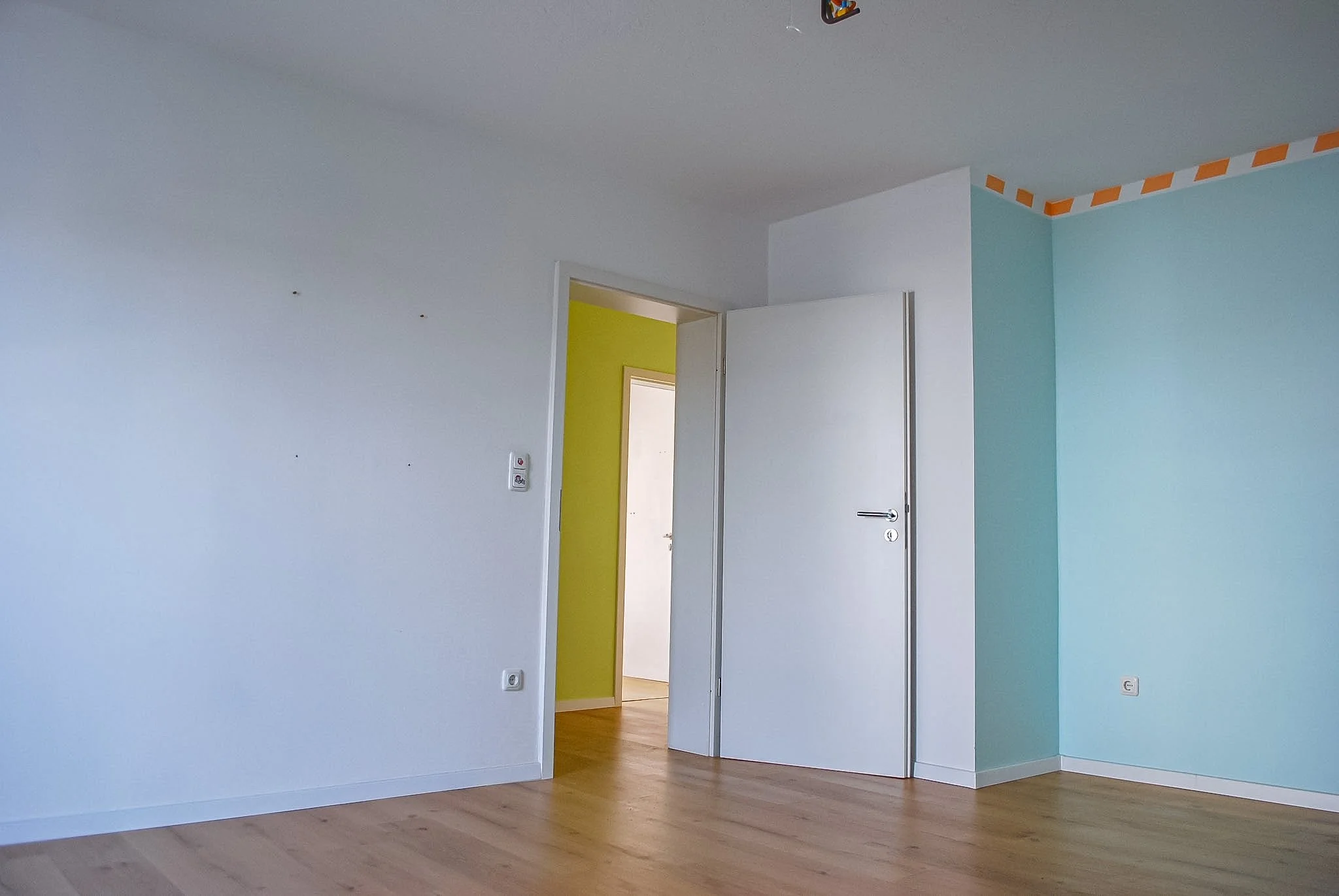 Ein Raum mit weißen und türkisfarbenen Wänden, einem offenen weißen Tür und Holzboden, mit sichtbarer angrenzender Raum mit gelber Wand.