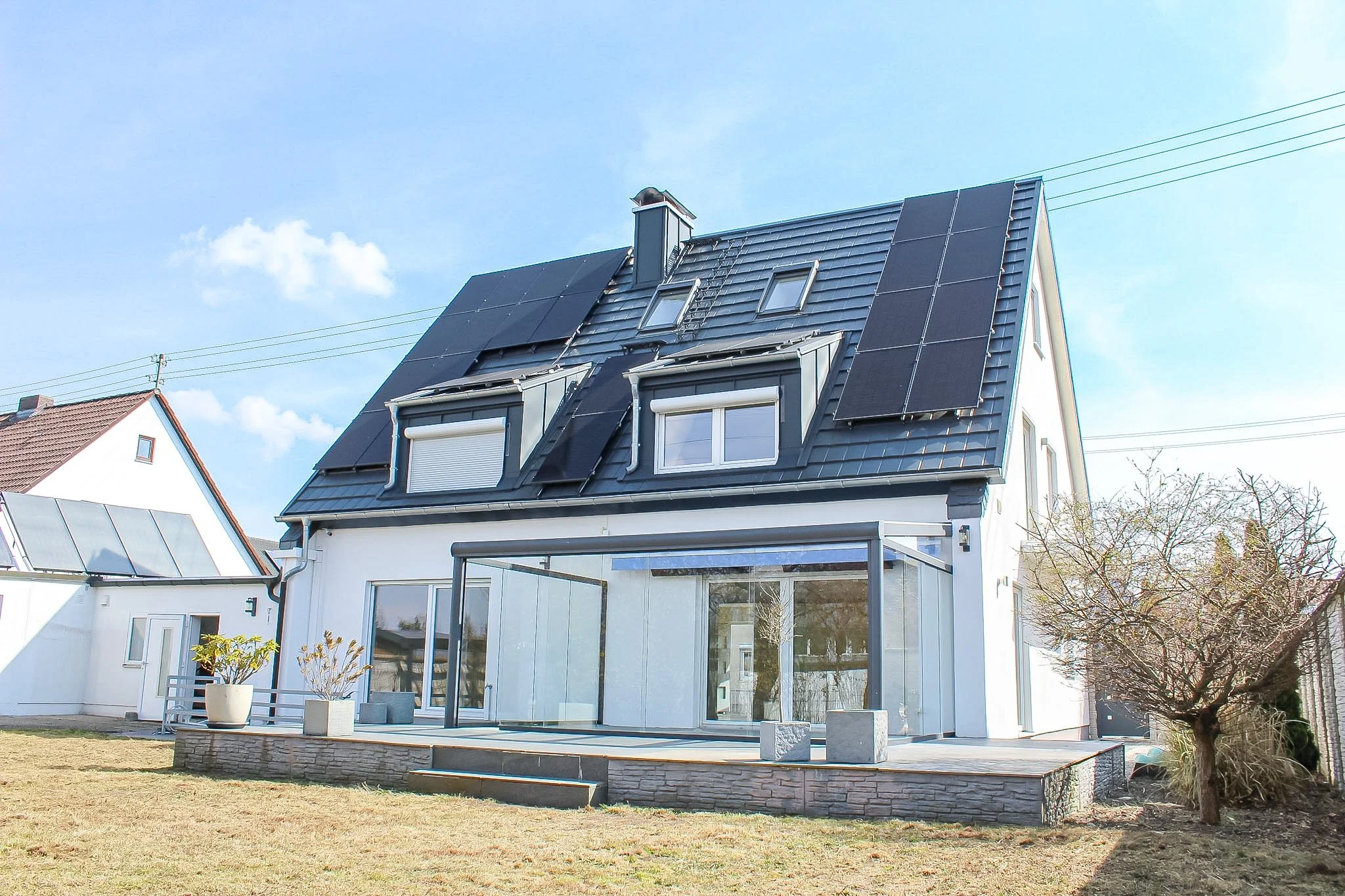 Modernes Haus mit Solaranlage auf dem Dach, großer Glasveranda, Garten mit Rasen und Balkon. Klarer Himmel mit wenigen Wolken.