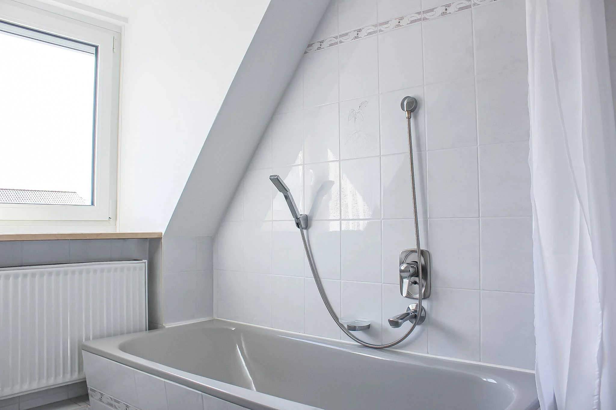 Ein Badezimmer mit einer Badewanne, einer Dusche mit Handbrause, weißen Wandfliesen, einem Fenster und einem Heizkörper.