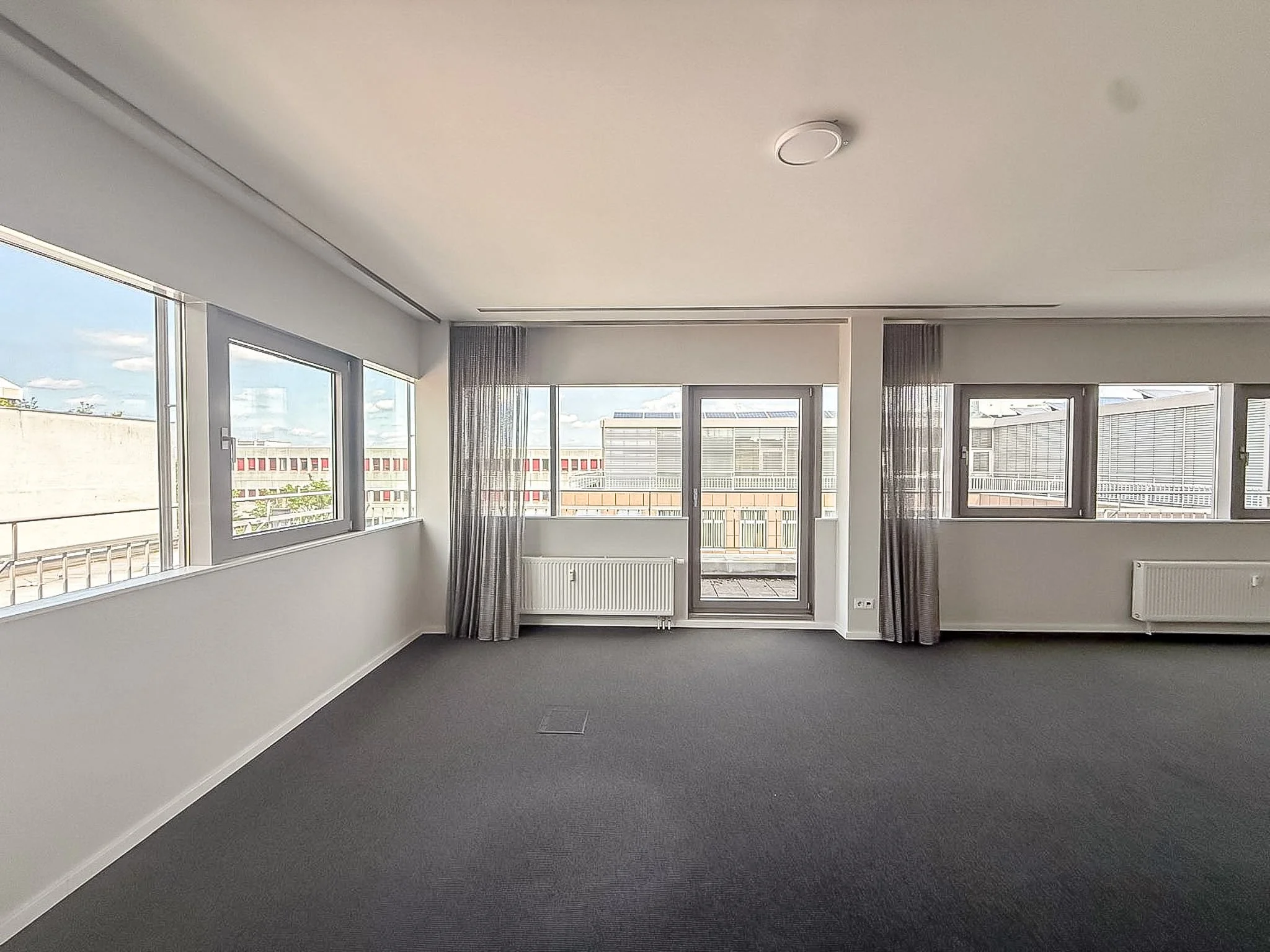 Leeres Zimmer mit großen Fenstern, dunklem Teppichboden und weißen Wänden