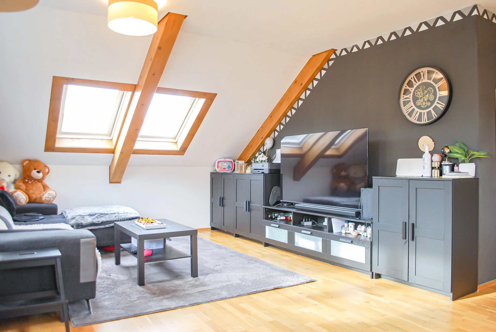 Ein Wohnzimmer mit einer schwarzen Couch, mehreren Teddy-Bären, einem Couchtisch, einer grauen Kommode mit Fernseher und Dekorationen, zwei Dachfenster, einer Uhr an der Wand und Holzbalken an der Decke.
